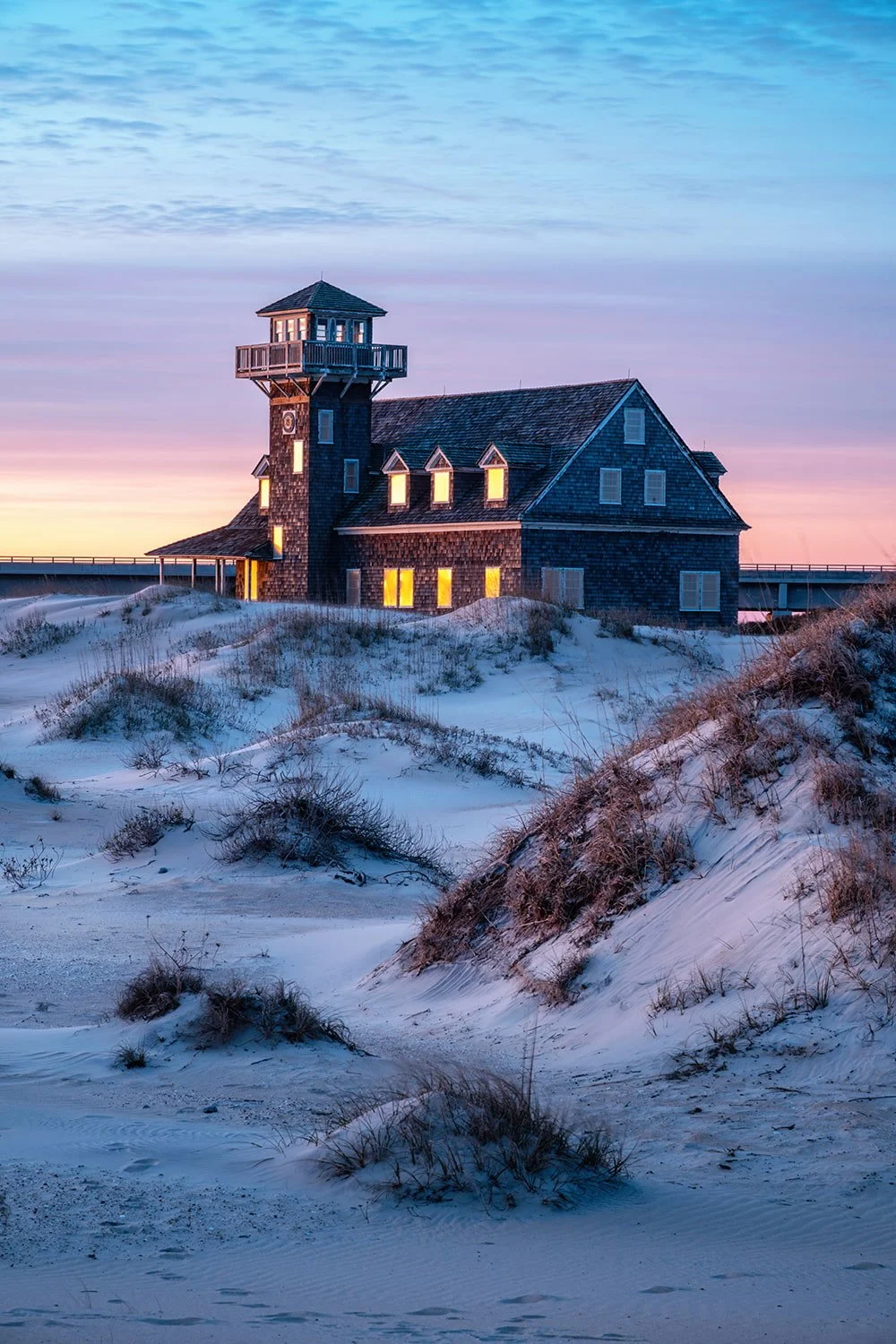 Oregon Inlet Life Saving Station at sunrise with sand dunes and grasses in the foreground, glowing building, and purple-yellow clouds in a cool-toned sky.