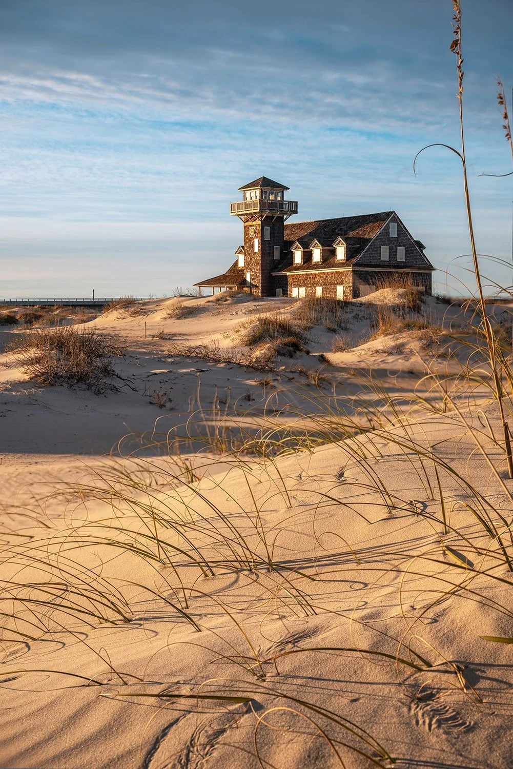 Oregon Inlet Life Saving Station at golden hour with sand and grasses in the foreground, dunes in the midground, building in the upper third, and sunlit front, under blue skies peeking through clouds.