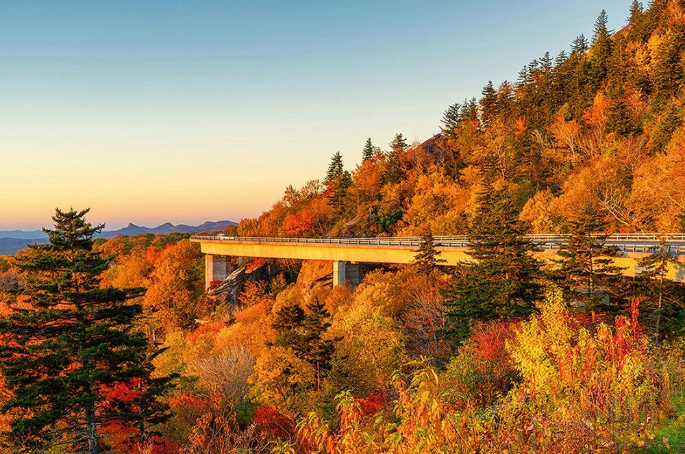 Linn Cove Viaduct in Western North Carolina in autumn with colorful foliage, illuminated by the rising sun under a cloudless sky, with a car in the distance showing motion blur on the rear section of the structure.