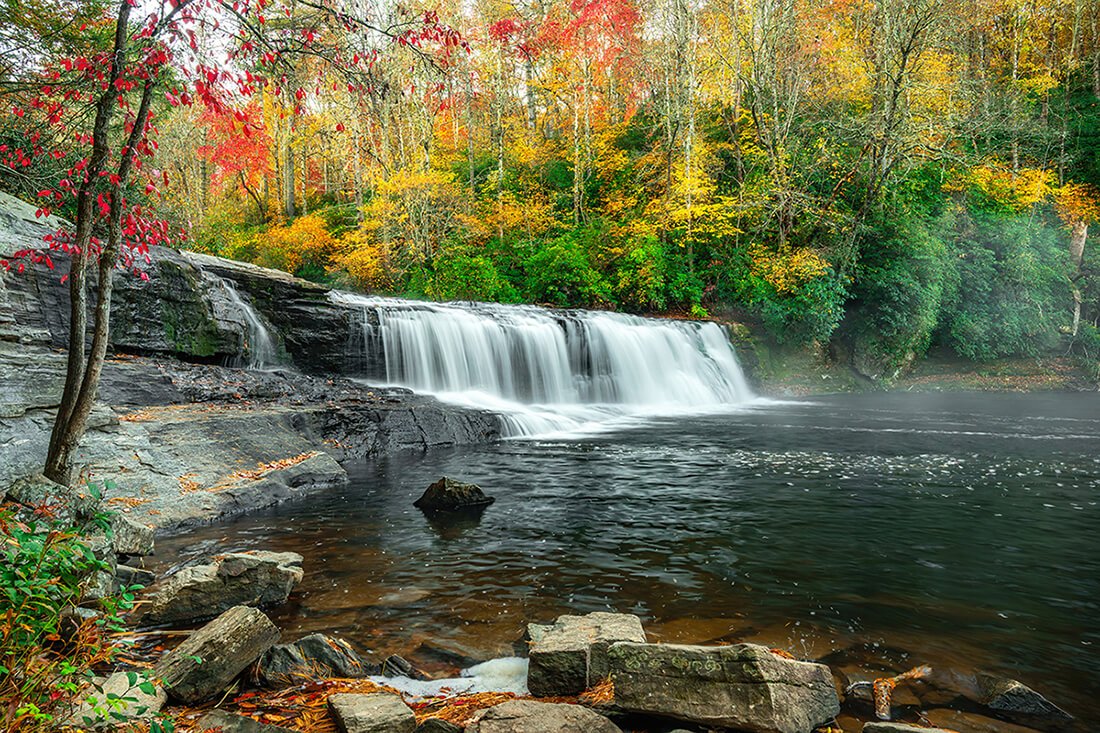Hooker Falls in autumn with vibrant fall foliage, rocks in the foreground, a lagoon in the middle, and the waterfall in the background.