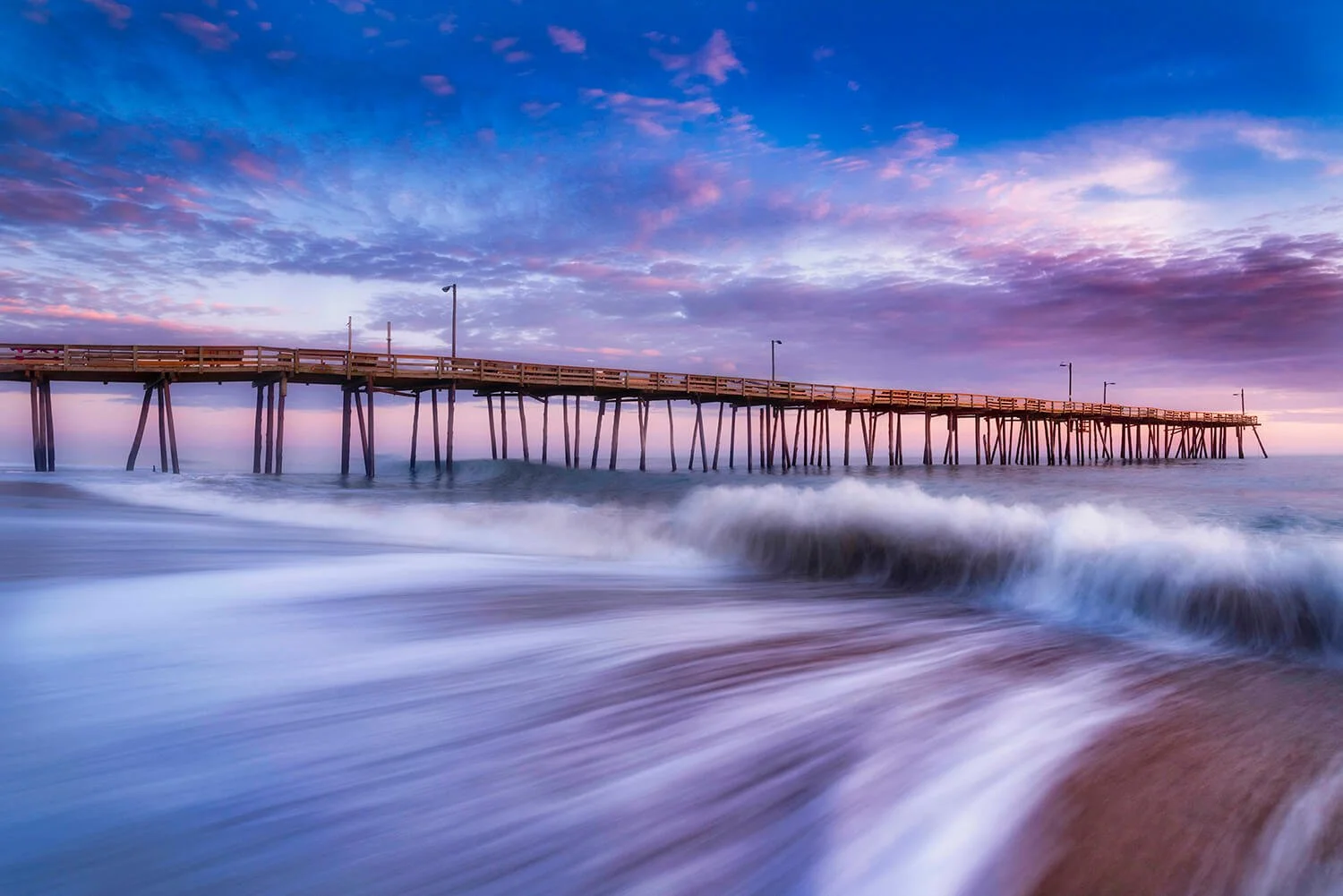 Nags Head fishing pier just after sunrise with flowing water in the foreground leading to a crashing wave beneath a deep blue sky with pink and purple overtones.
