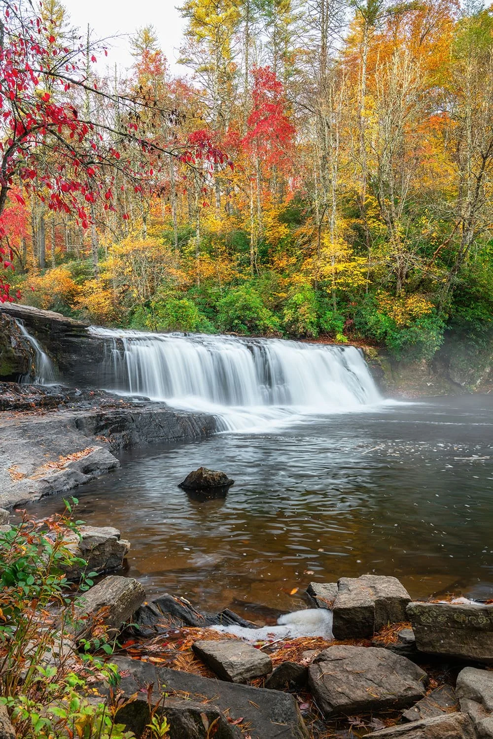 Hooker Falls at Dupont State Recreational Forest, in Brevard, North Carolina during autumn with vibrant fall foliage, rocks in the foreground, a lagoon in the middle, and the waterfall in the background.