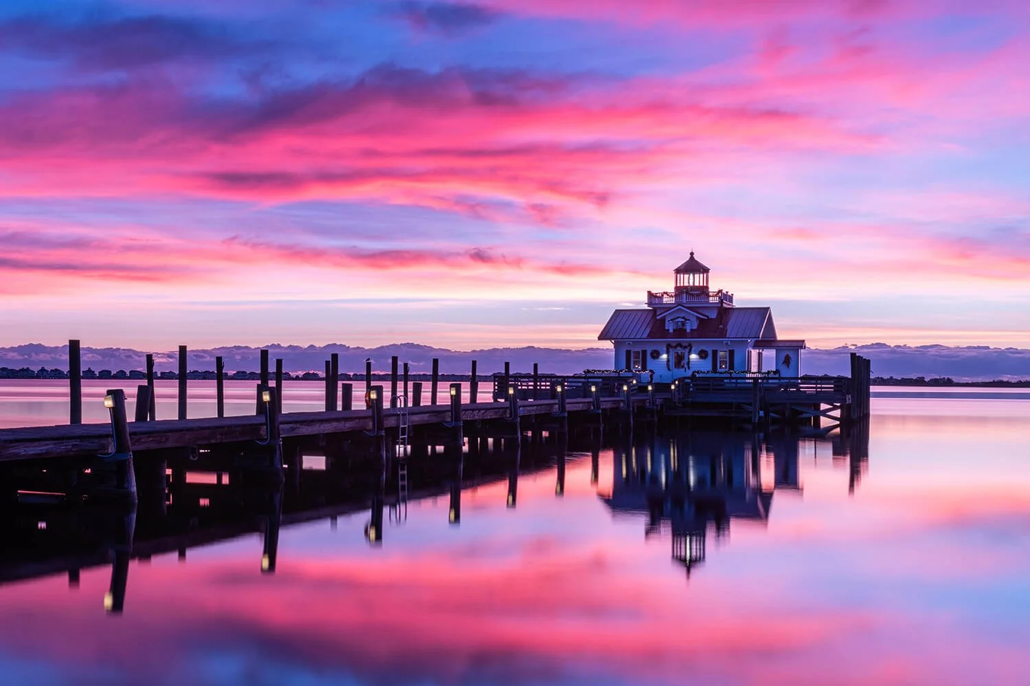 Roanoke Marshes Lighthouse in Manteo, North Carolina at sunrise, viewed from a 45-degree side angle with a dock leading to the building, pink and blue sky reflecting in the water, dock lights on, and holiday decorations including a wreath and garland