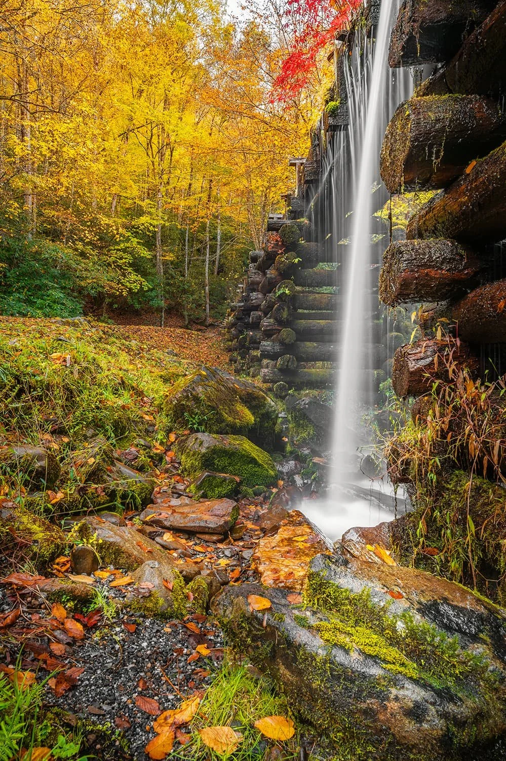 Water flowing over the wooden flume at Mingus Mill in the Great Smoky Mountains National Park with rocks in the foreground, water hitting the ground, and vibrant fall foliage in the background.