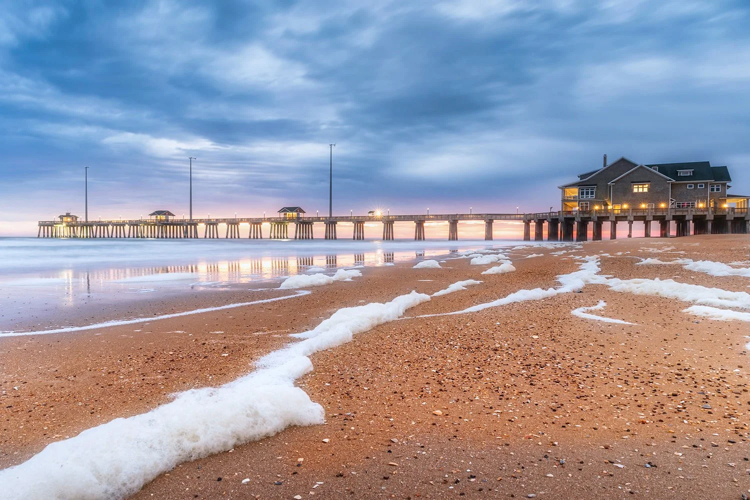 Jennette’s Pier in Nags Head, North Carolina at sunrise at low tide, with reflections in the water, seafoam and shell debris in the foreground, and the sun breaking a moody cloudy sky.