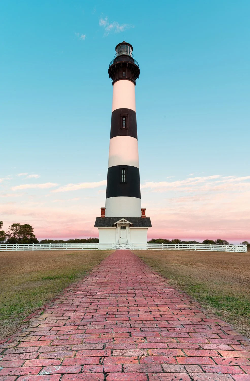 Bodie Island Lighthouse just after sunrise with a brick walkway leading toward the building, captured from a low angle in clean morning light.