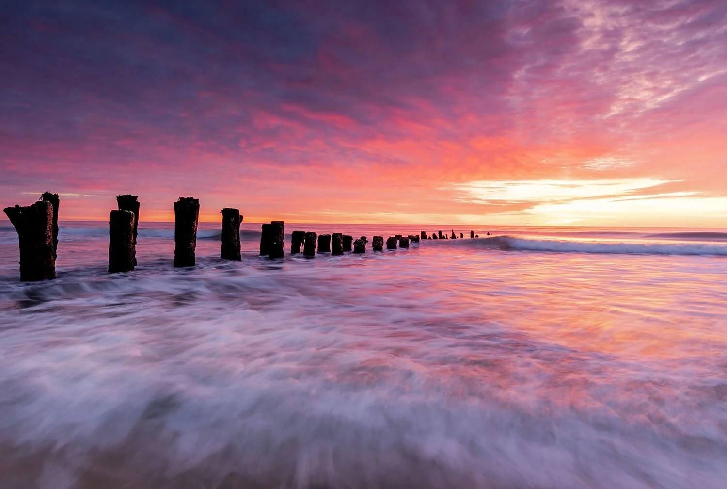 Carolina Beach at sunrise with water motion in the foreground, silhouetted piles extending from the foreground to mid-frame on the left, catching the light of the rising sun.