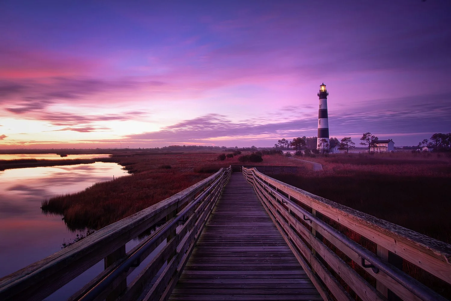 Bodie Island Lighthouse in North Carolina at sunrise with a walkway leading from the observation deck, lighthouse positioned to the right, and purple overtones in the sky.