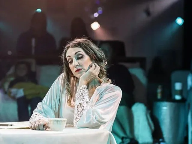 Emily sits pensively on stage, glancing down at a paper and pen sitting next to a teacup on a small table. Stage lights and an audience are blurred in the dark background.