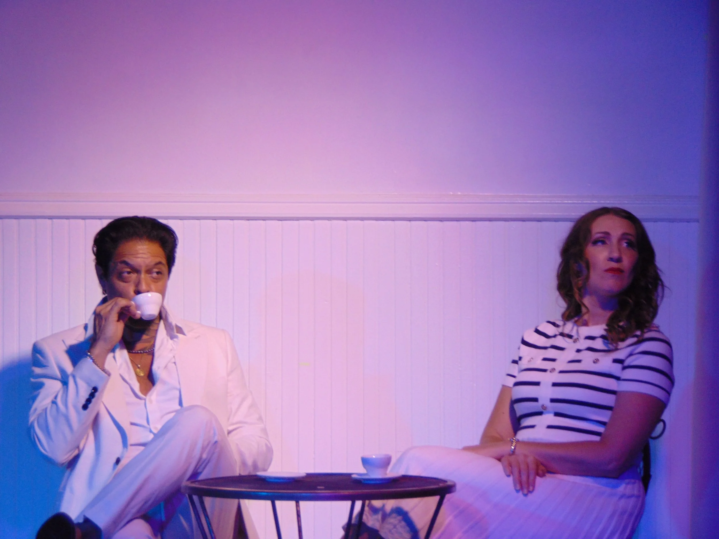 Two actors sit at a small patio table, one sipping espresso, in Robbie, an immersive theater show in Eureka Springs Arkansas near Fayetteville, Bentonville, Rogers, and Springdale.