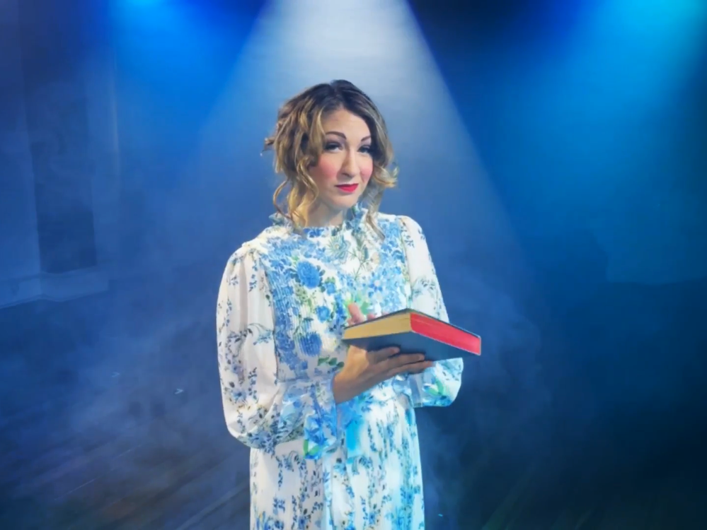 An actor in a white dress adorned with blue flowers holds a book in I Haunt You, an immersive theater show in Eureka Springs Arkansas, near Bentonville, Fayetteville, Springdale, and Rogers.