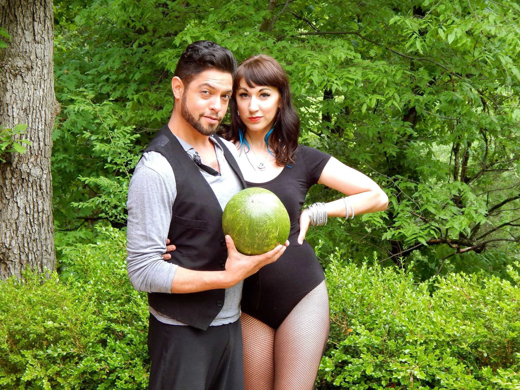 Raymond looks at the camera holding a watermelon while Emily stands next to him with an arm around his waist in front of a lush Ozark background