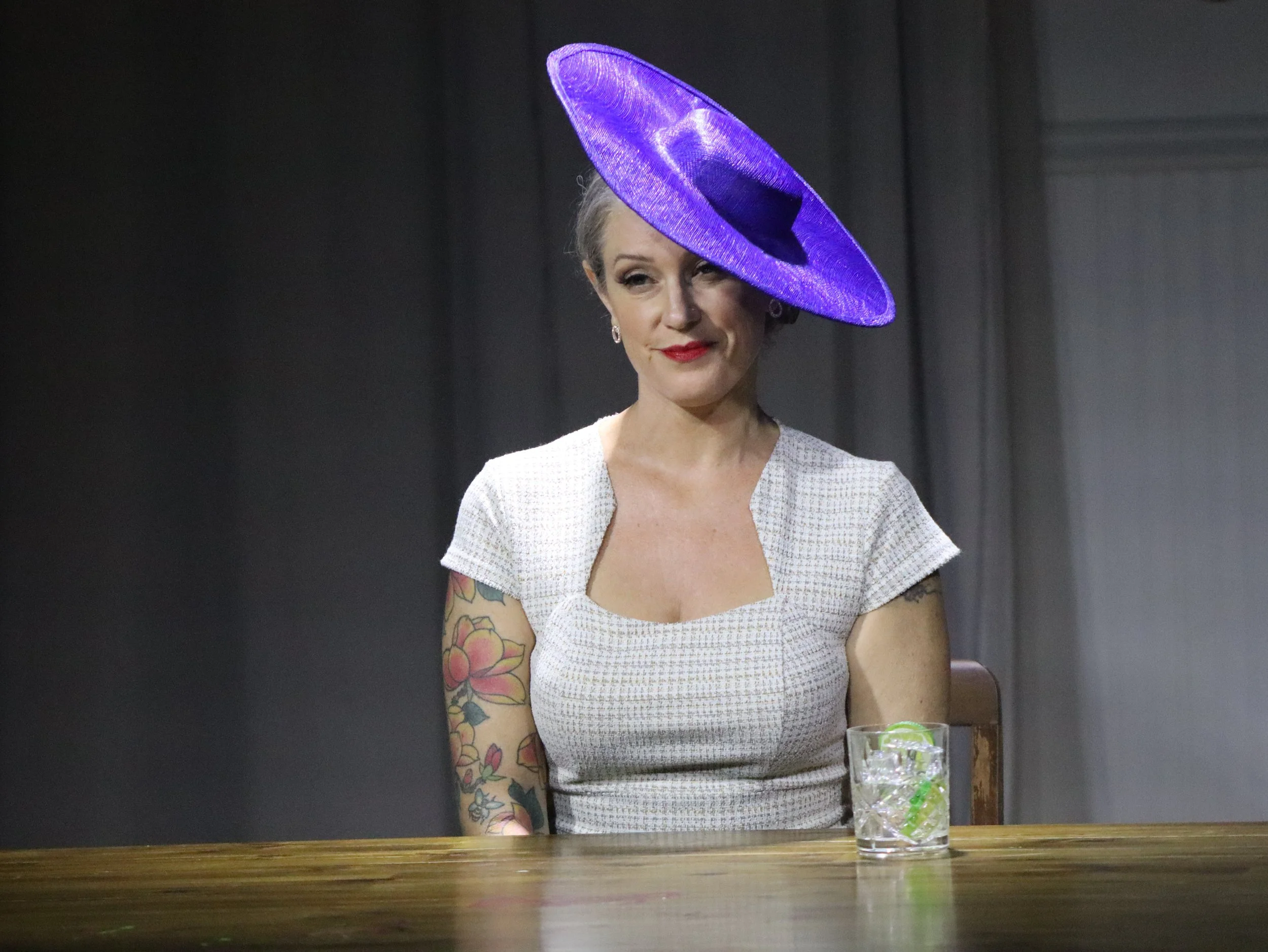An actor in a fitted white dress and purple hat giving a sly smile in Robbie, an immersive theater show in Eureka Springs Arkansas, near Fayetteville, Bentonville, Rogers, and Springdale.