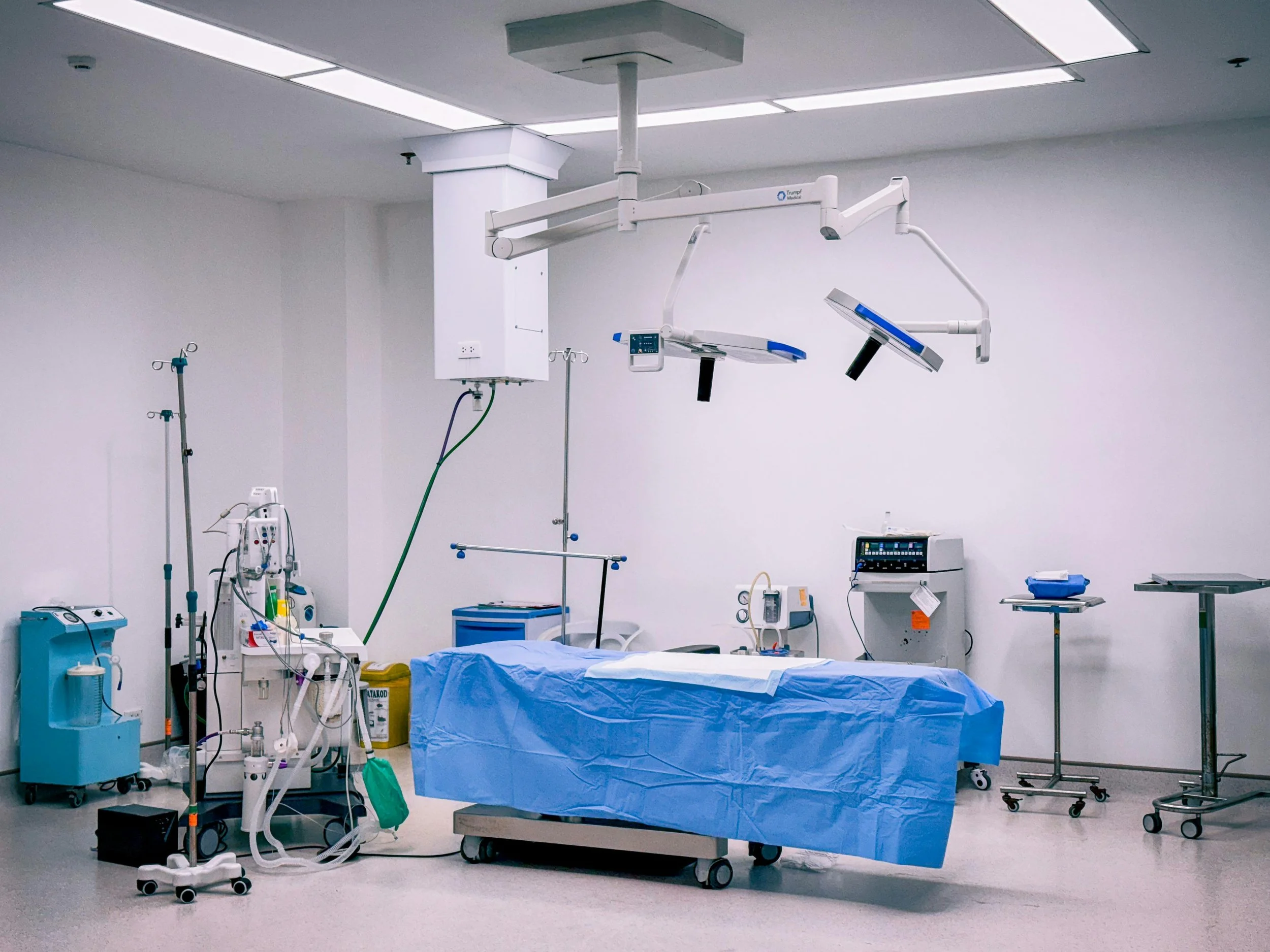 Empty hospital operating room with surgical table covered in blue drape, surgical lights overhead, and various medical equipment and monitors around the room.