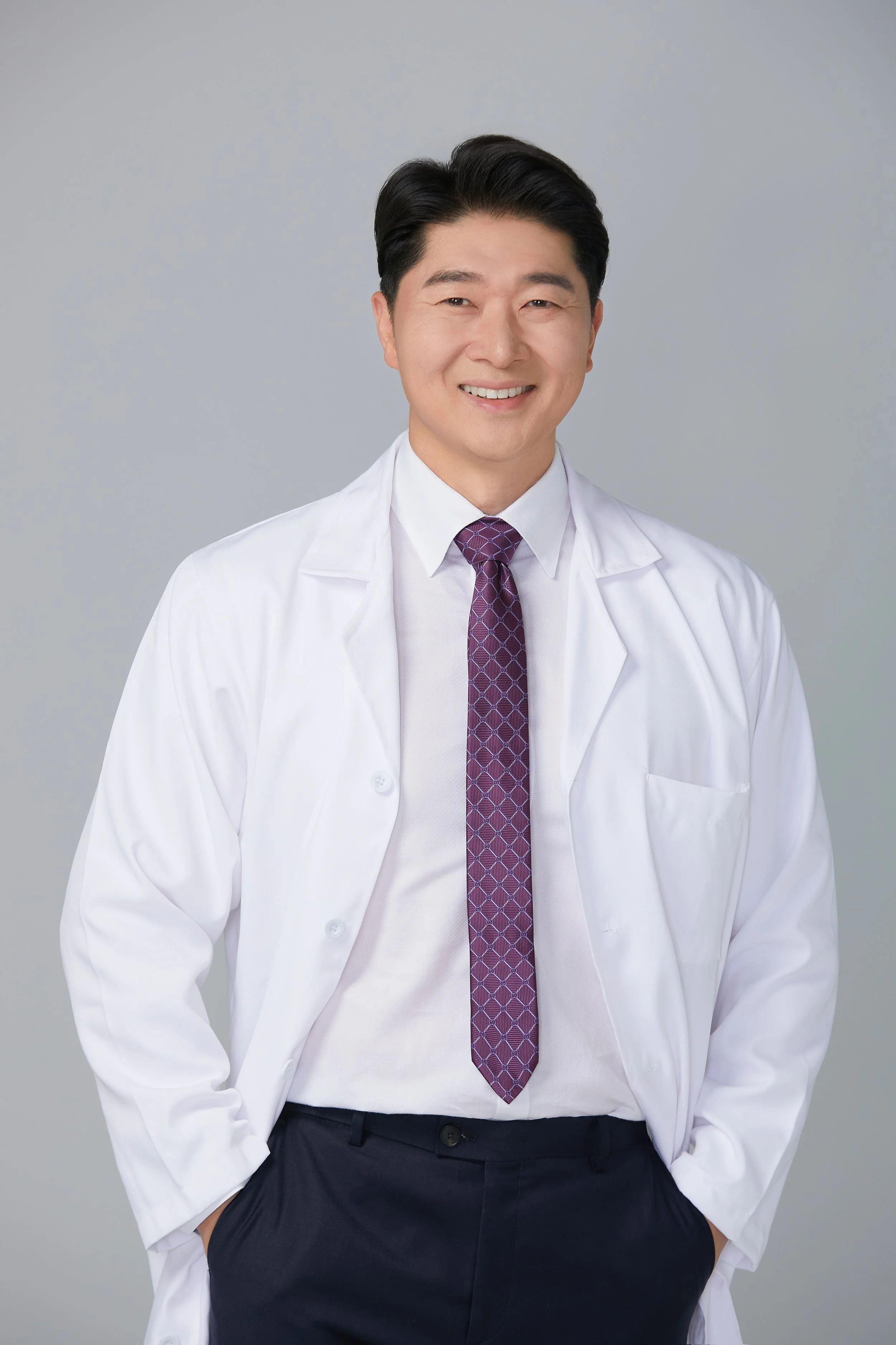 Smiling male doctor in white coat and purple patterned tie, standing with hands in pockets against light gray background.