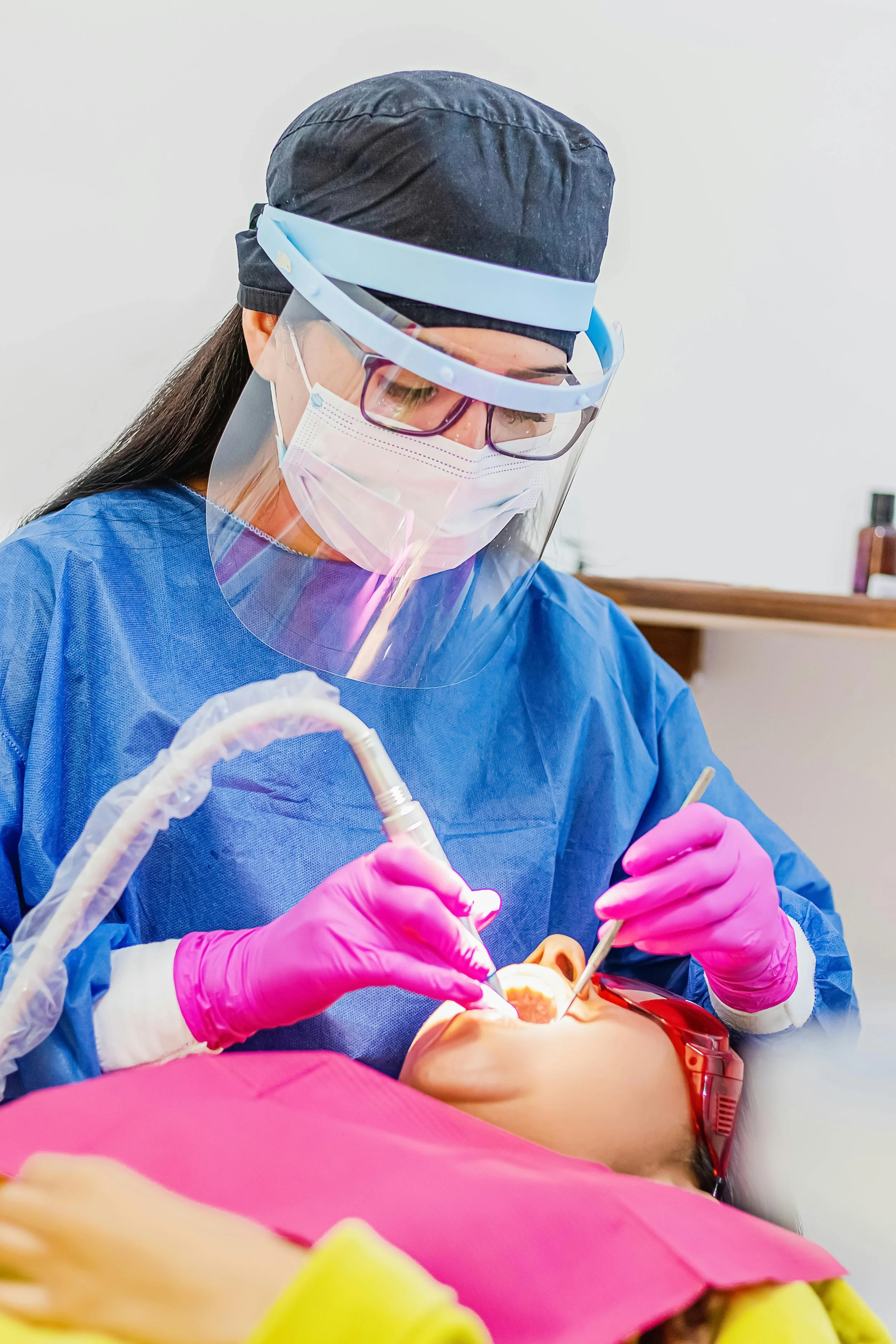 A dentist in protective gear, including a face shield, mask, and gloves, working on a patient's mouth in a dental office.