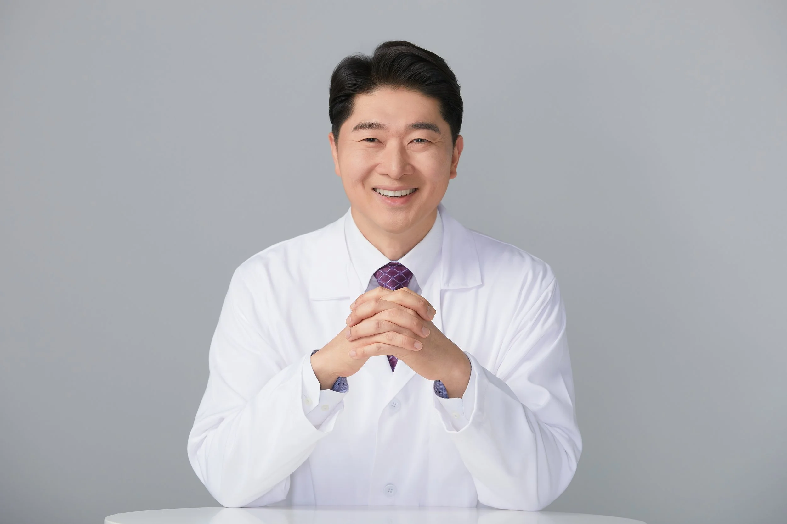 A smiling male doctor wearing a white lab coat and a purple tie, sitting at a white table with hands clasped, against a plain gray background.