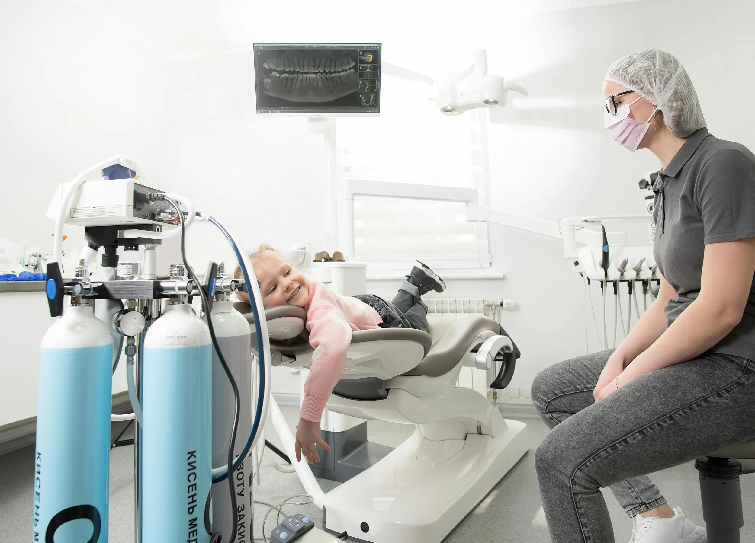 A young girl lying in a dental chair, smiling at a female dental professional wearing a mask and glasses in a bright dental office with a panoramic dental X-ray displayed on a monitor.