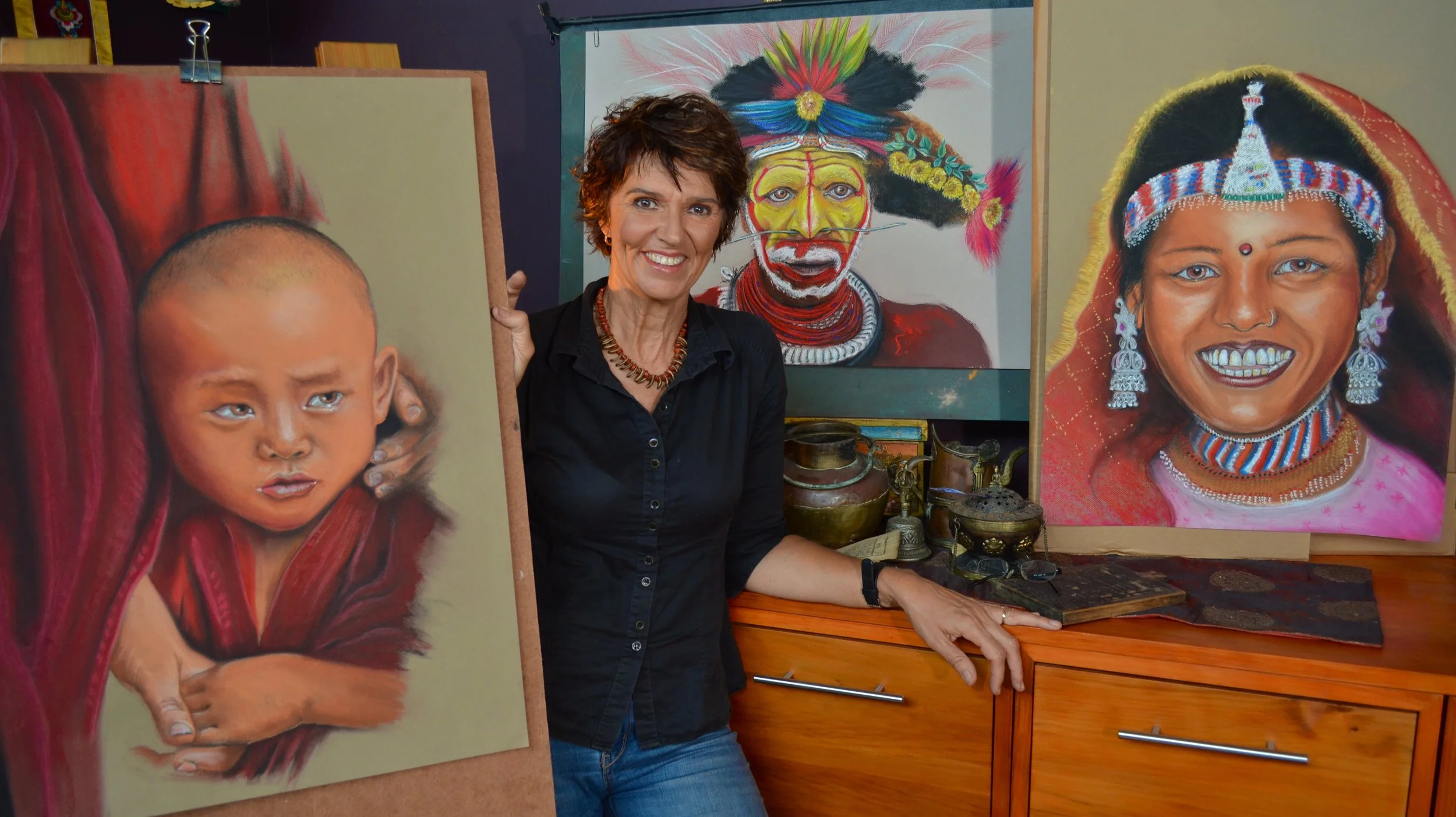 An art gallery or studio with a woman standing between two large portrait paintings of children wearing traditional attire. Different artwork styles display expressive faces and vibrant colors.