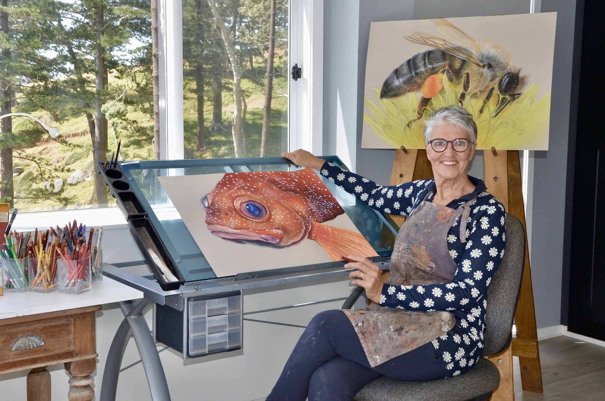 Sorrel Wilby in her art studio. She is smiling as she works on a large painting of a fish with vibrant colors. The studio has a window showing trees outside, and on the wall behind her is a painting of a bee on a yellow flower.