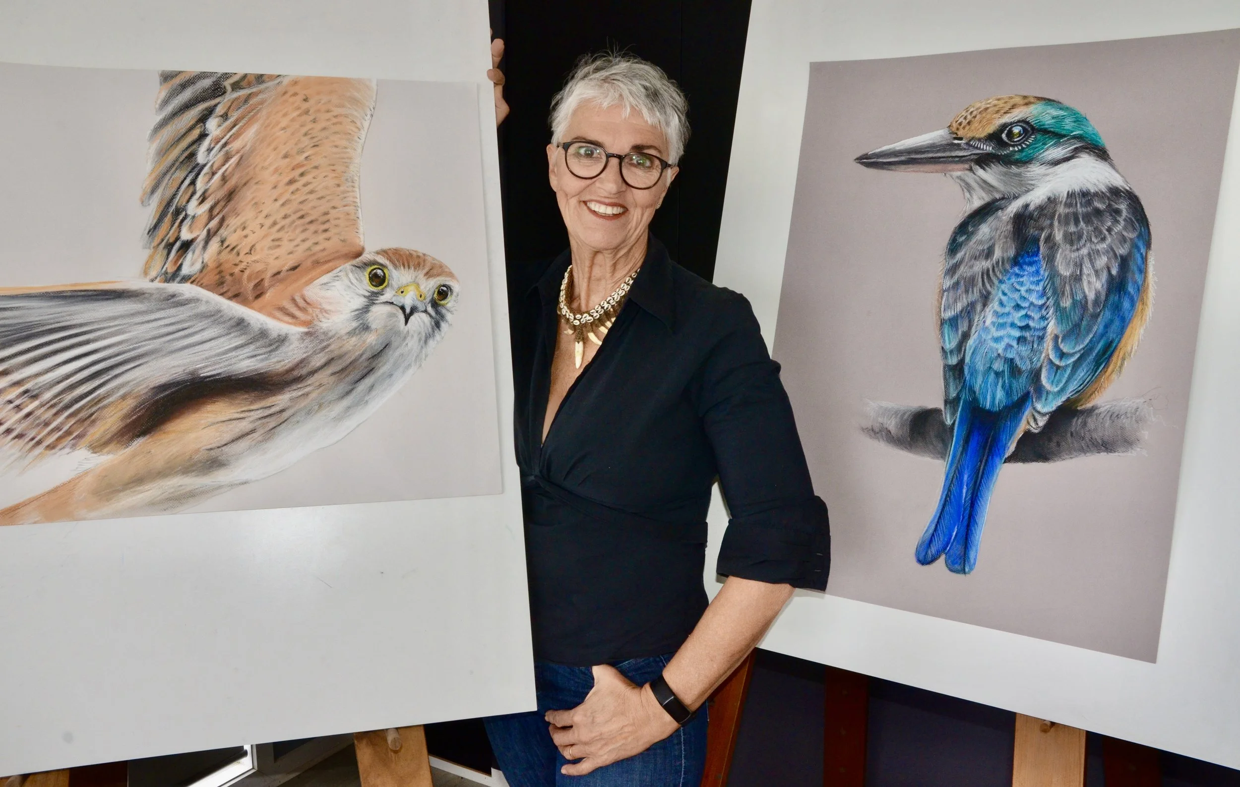 Sorrel Wilby, wearing a black shirt and necklace, standing between two large, detailed paintings of colorful birds, smiling at the camera.