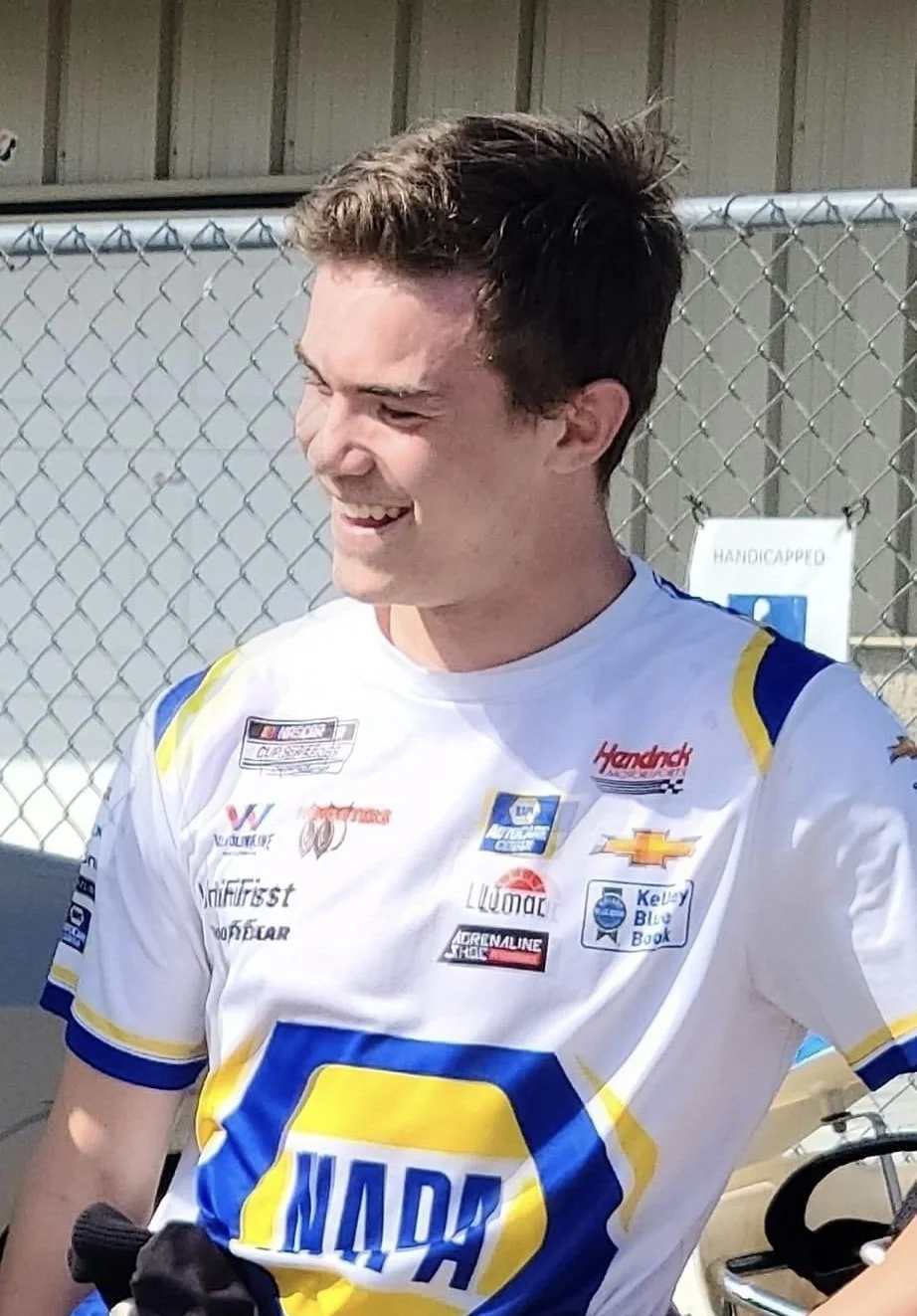 A young man smiling, wearing a racing jersey with multiple sponsor logos, standing in front of a chain-link fence.