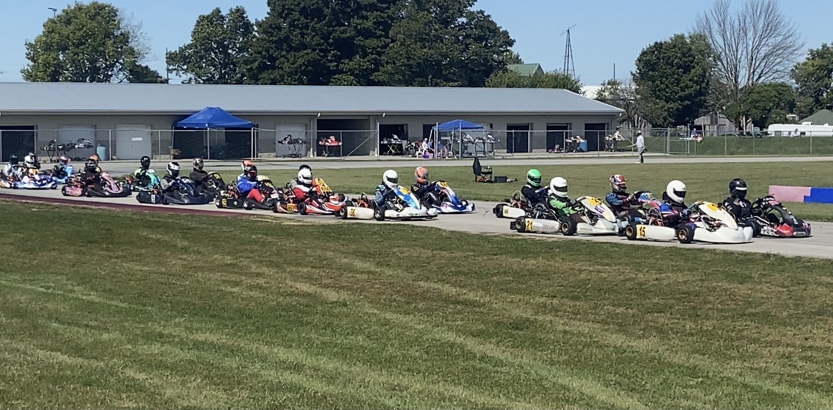 A line of go-karts with drivers wearing helmets on a race track, with a grassy area in the foreground and a building with blue tents and spectators in the background.
