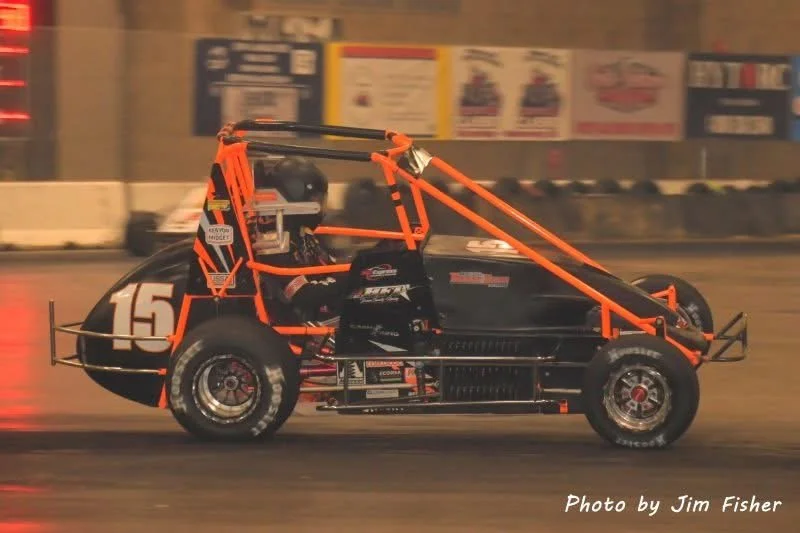 A black and orange off-road race car with the number 15 on the side, racing on a dirt track at night, driven by a helmeted driver. The background has blurred advertisements.