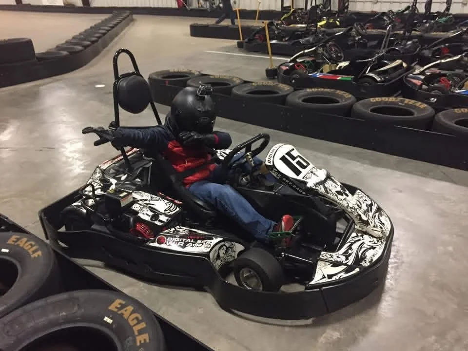 Person wearing a black helmet and red jacket driving a go-kart with black and white graphics inside an indoor racing facility, with tires and other go-karts in the background.
