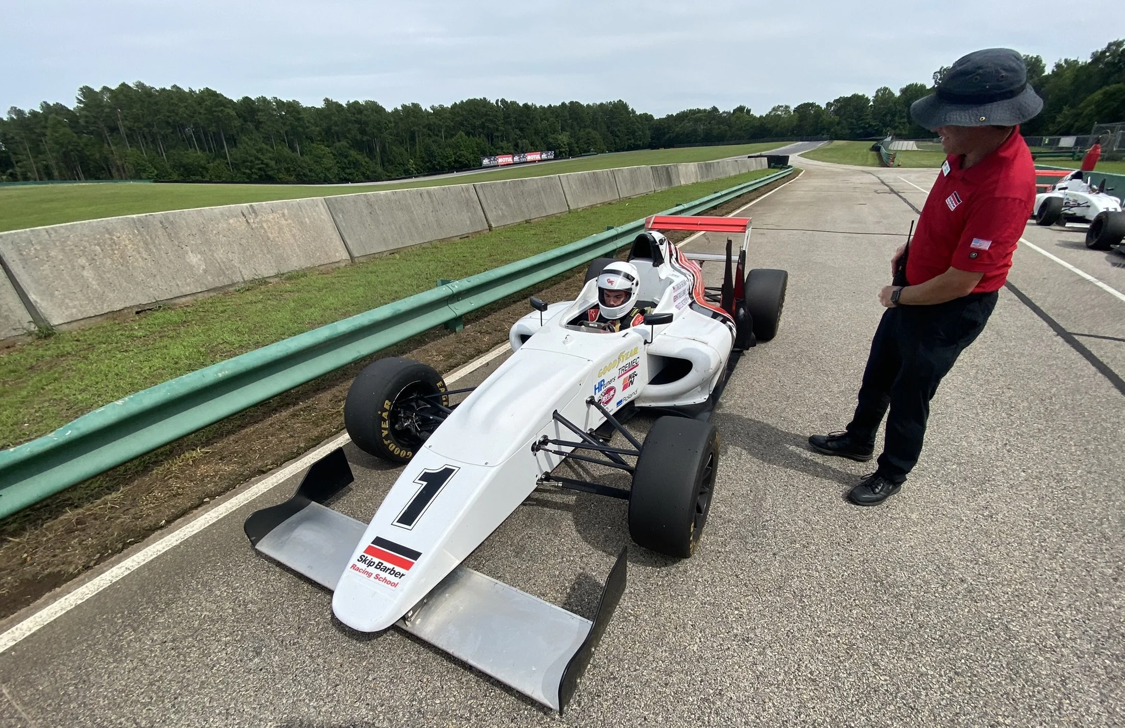 A race car with the number 1 on it parked on the track, with a person in a white helmet in the driver's seat and a man in a red shirt and black hat talking to the driver on the side of the car.