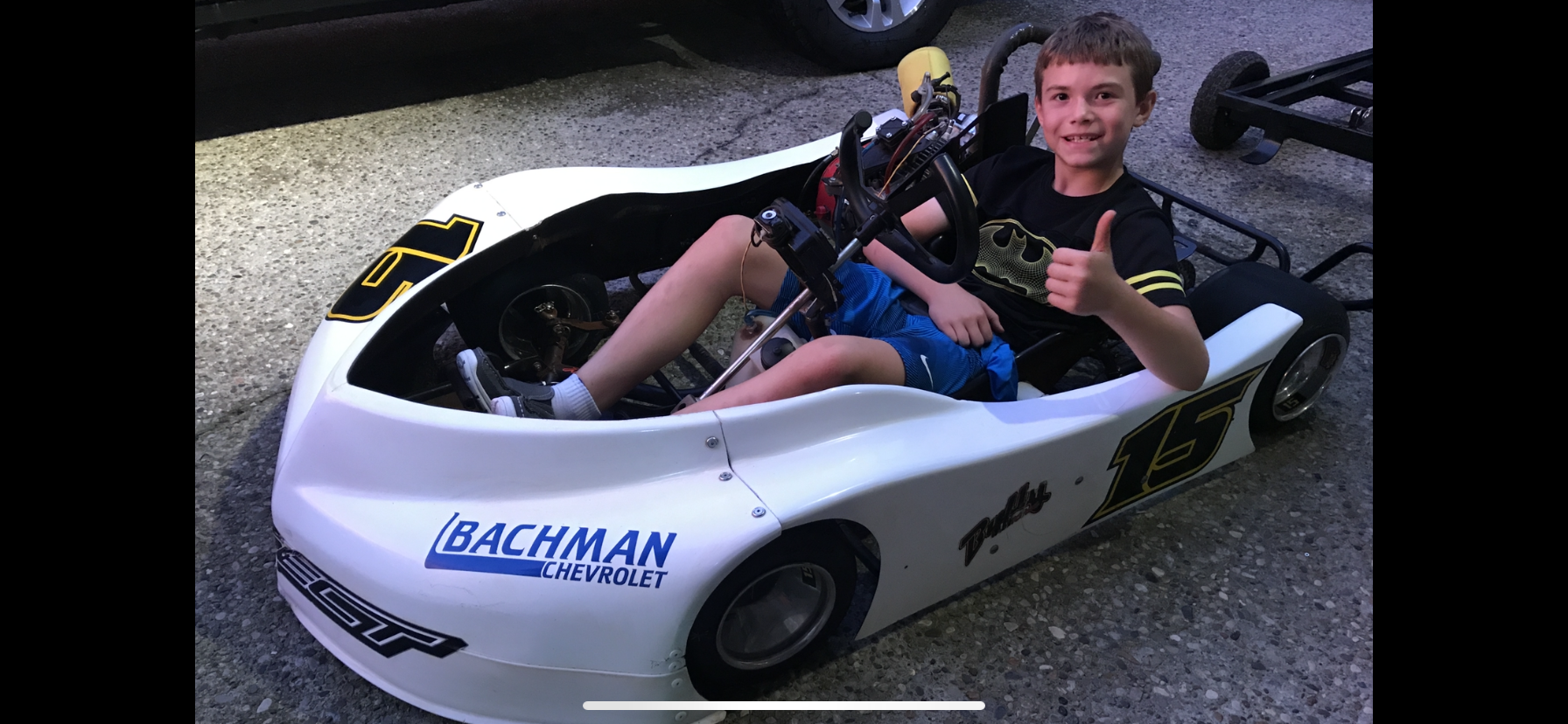 A young boy sitting in a white go-kart with the number 15, giving a thumbs-up, wearing a black Batman t-shirt and blue shorts, on a gravel surface.