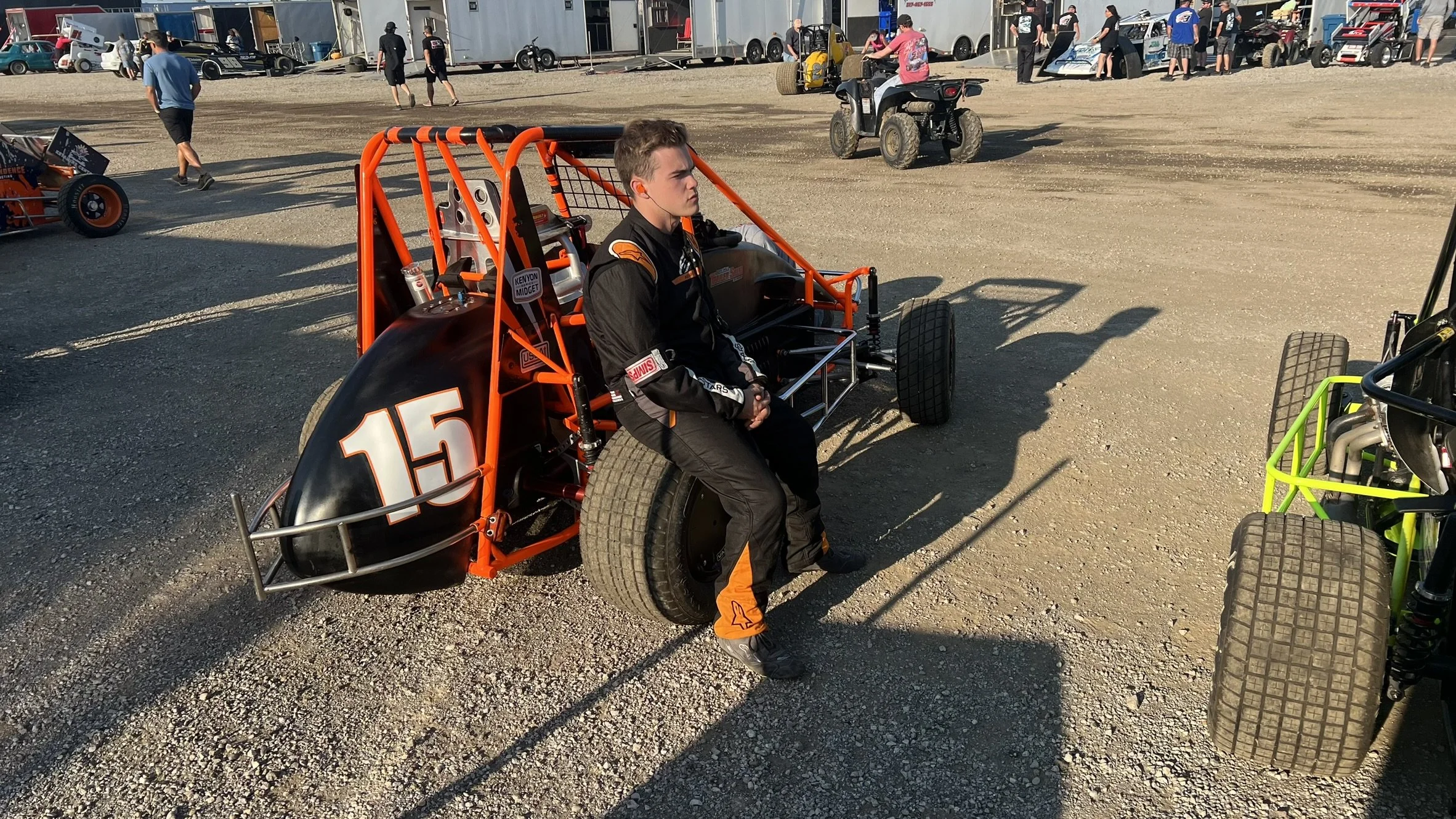 A young man dressed in a black racing suit with orange accents sitting on the side of a black and orange dirt race car number 15, in a dirt lot with other race cars, people, and trailers in the background.