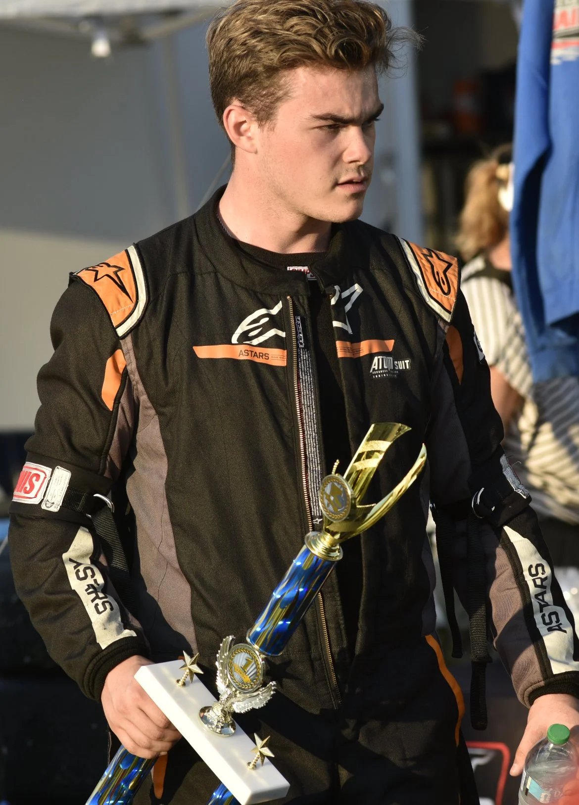 A young man in racing overalls holding a trophy with a serious expression, standing outdoors.