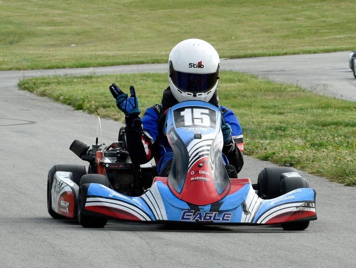 Go-kart driver wearing a white helmet with a blue racing suit and gloves, making a peace sign with hand, on a race track.