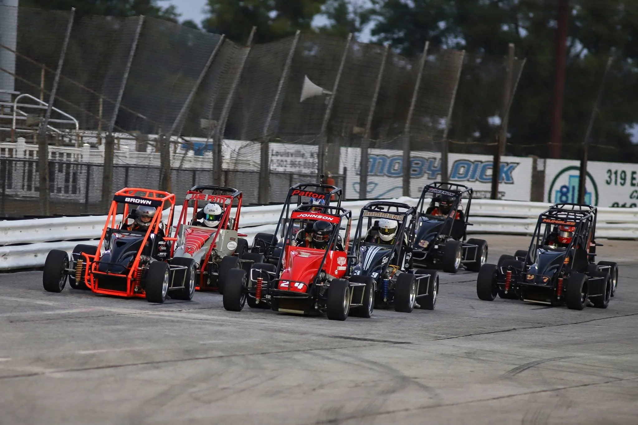 Six mini race cars on a track, with drivers wearing helmets, competing in a race at a dirt oval track with fencing and advertising banners in the background.