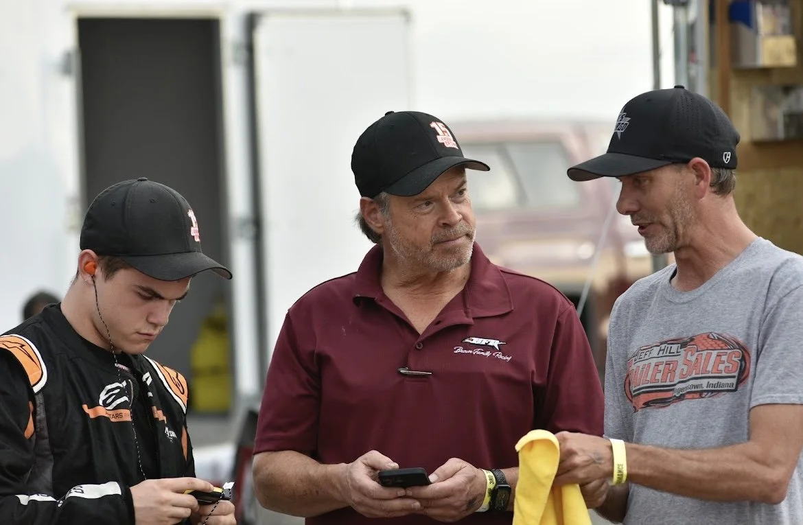 Three men having a conversation at a racing event, one young man in racing suit and two older men wearing caps, one holding a phone and the other a yellow cloth.
