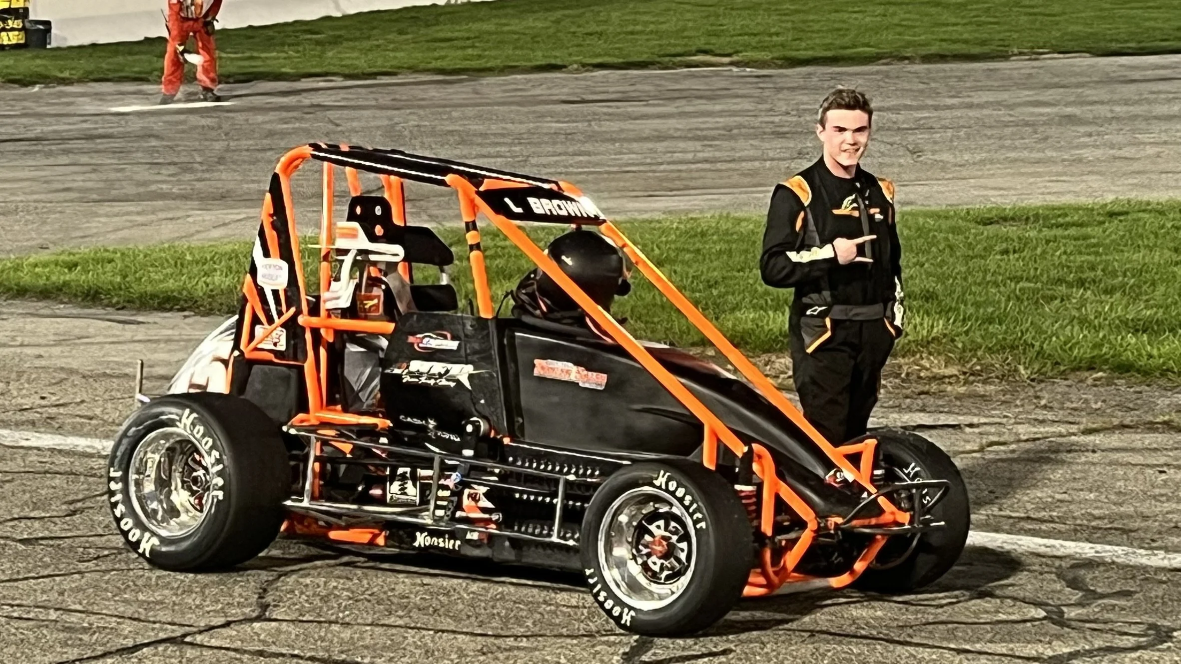 A race car driver in racing suit standing next to an orange and black race car on a track, with a grassy area in the background.