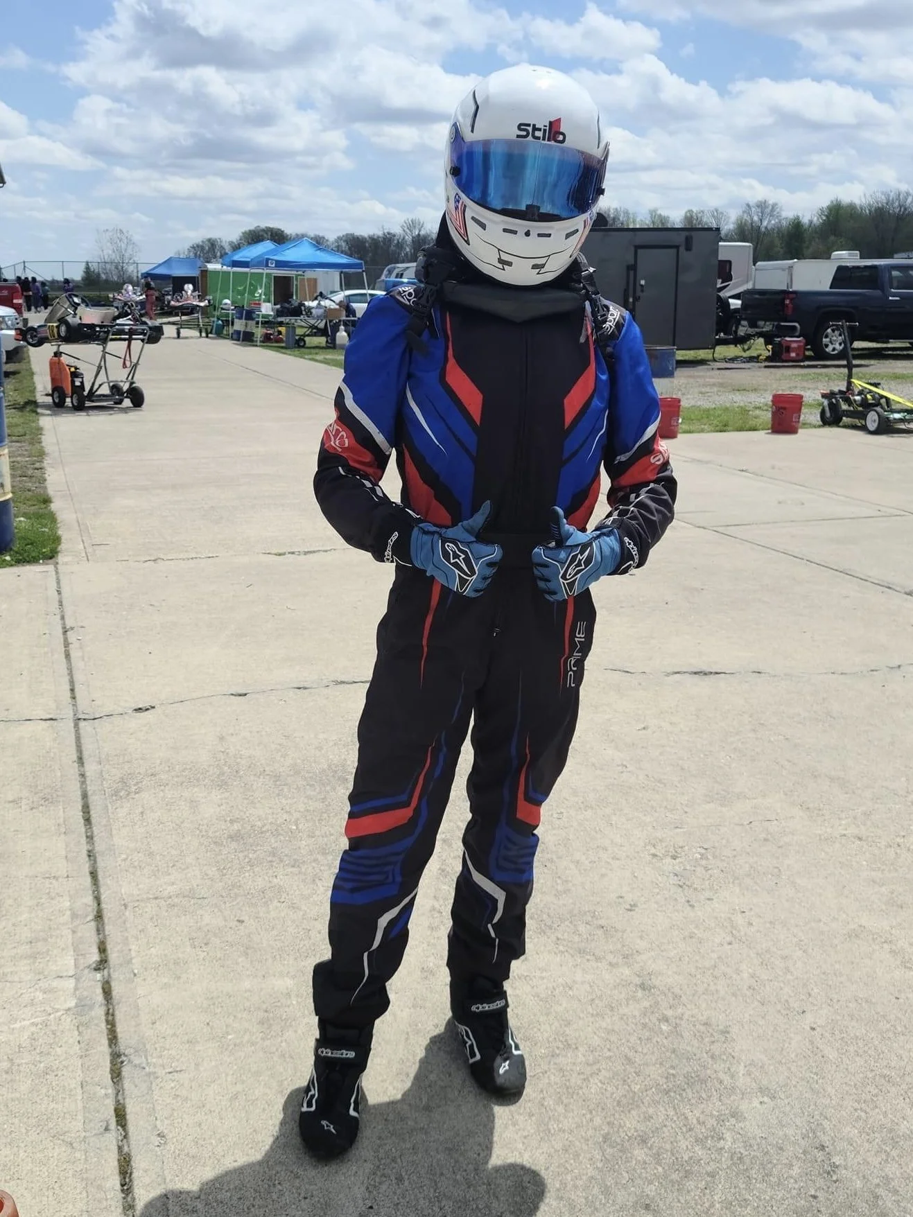 Person in racing suit and helmet standing on a paved area at a racing event.