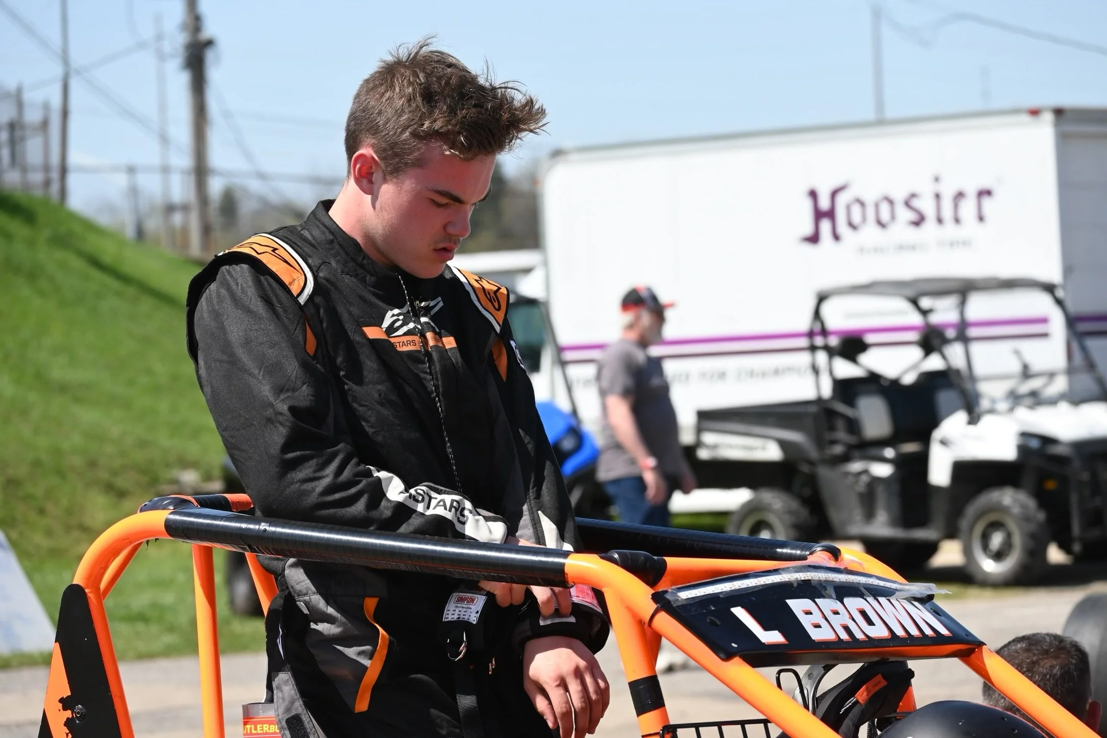 A young man in racing gear standing next to an orange race car with the name 'L BROWN' on it, looking down and adjusting his wrist strap, with a grassy hill, a man in a cap, and utility vehicles in the background on a clear day.