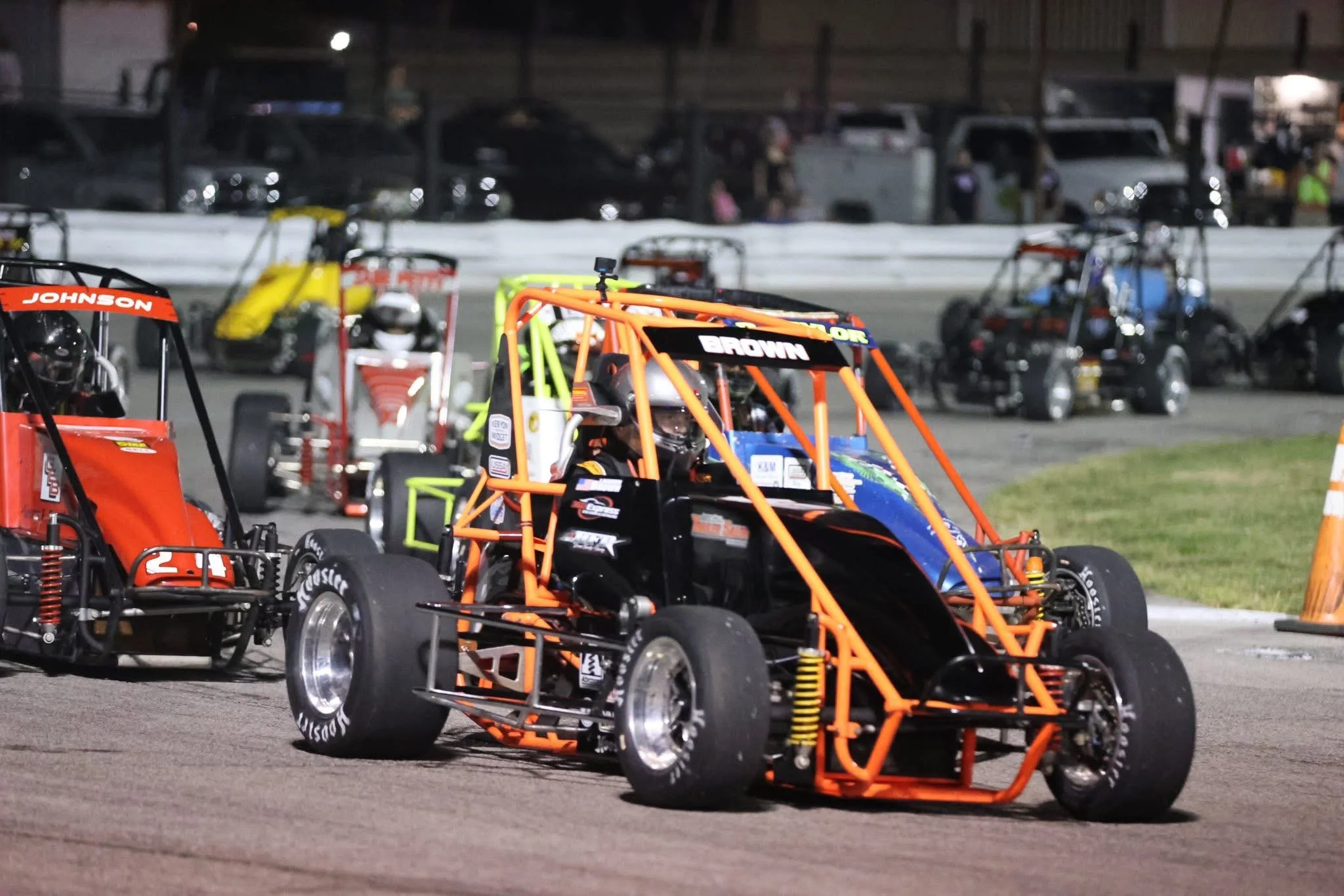 Nighttime race track with several small race cars parked on the asphalt, including a black and orange race car with a driver inside, orange roll cage and yellow suspension spring, other cars and vehicles in the background.
