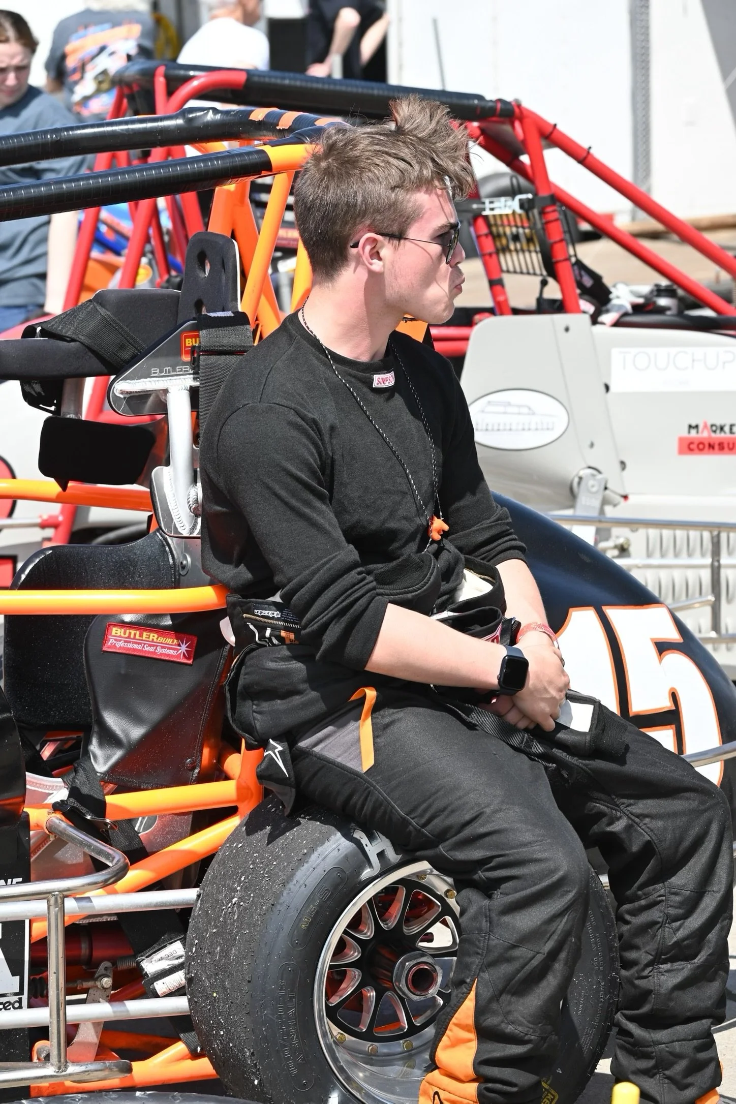 A young man in black racing gear and sunglasses sitting on a race car with the number 15, in a racing paddock with other race cars and people in the background.