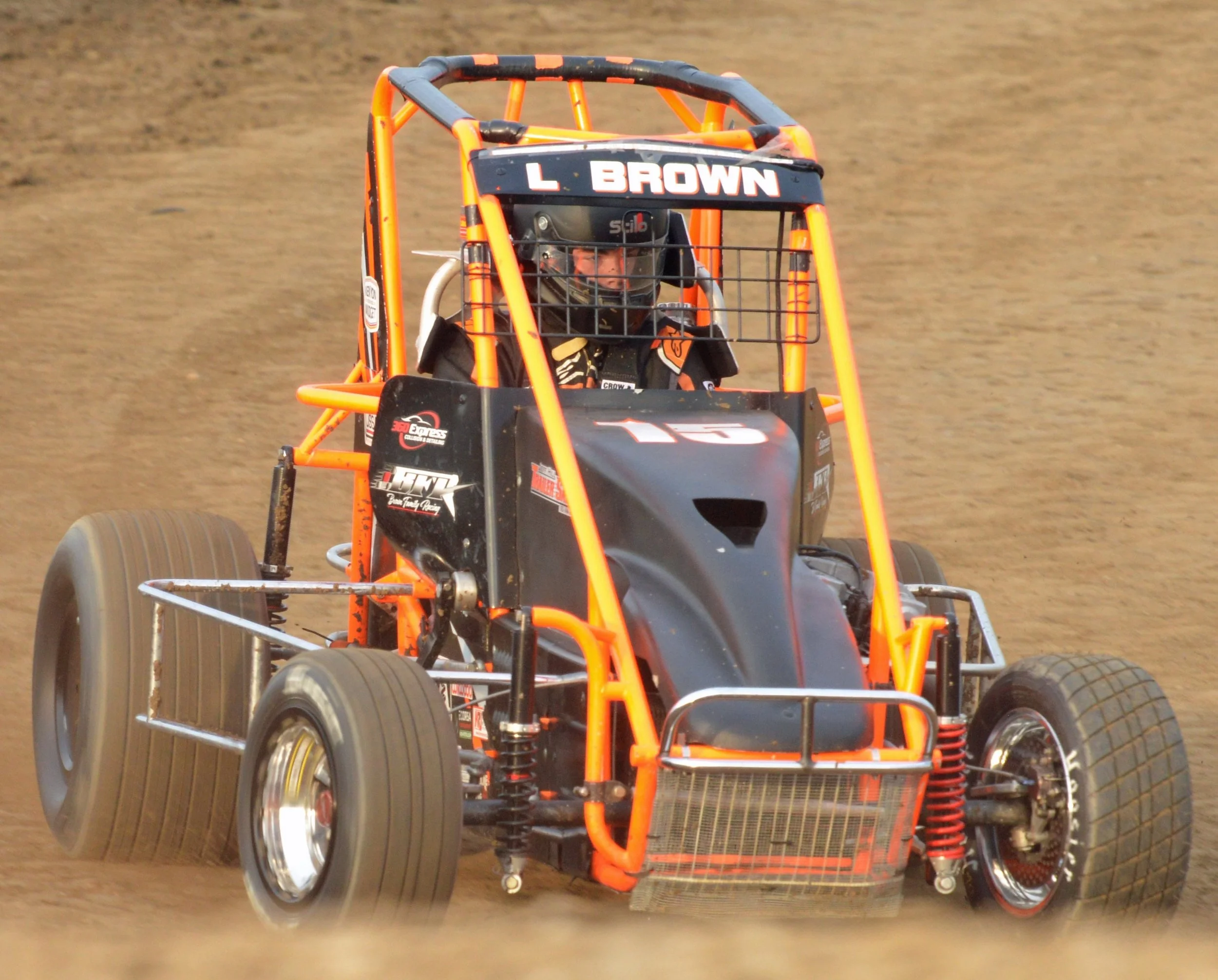 An orange and black dirt track racing car with a driver inside, wearing a helmet, driving on a dirt track.