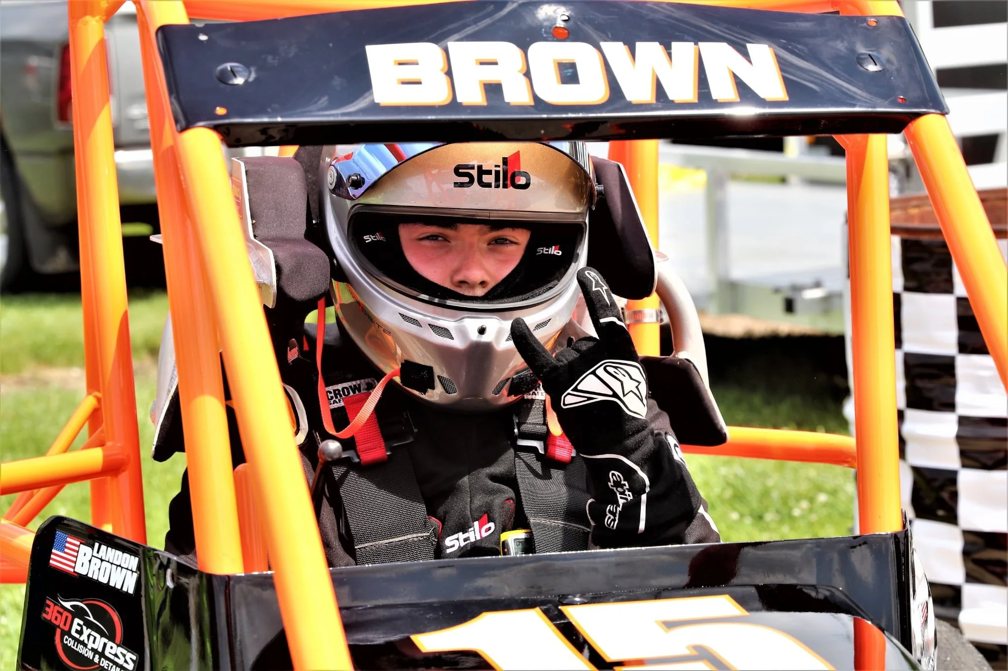A young race car driver wearing a silver helmet and black racing suit, sitting in a black and orange race car with the number 15, making an 'OK' hand gesture.