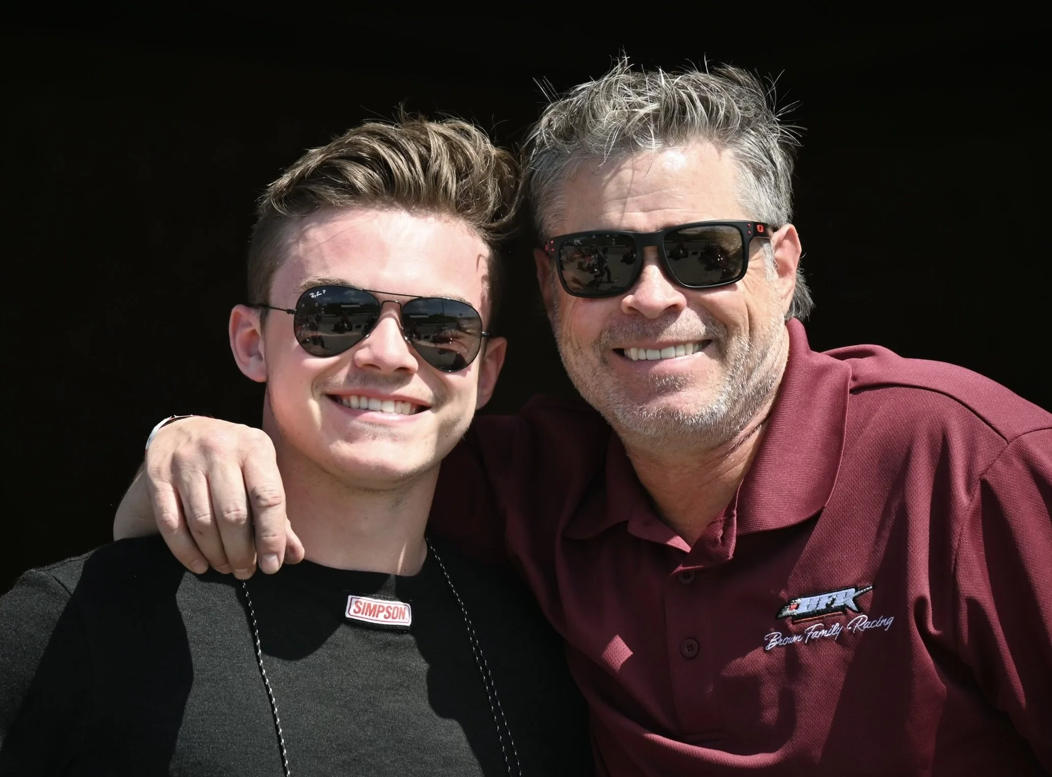 Two men smiling, wearing sunglasses, with their arms around each other, posing outdoors against a dark background.