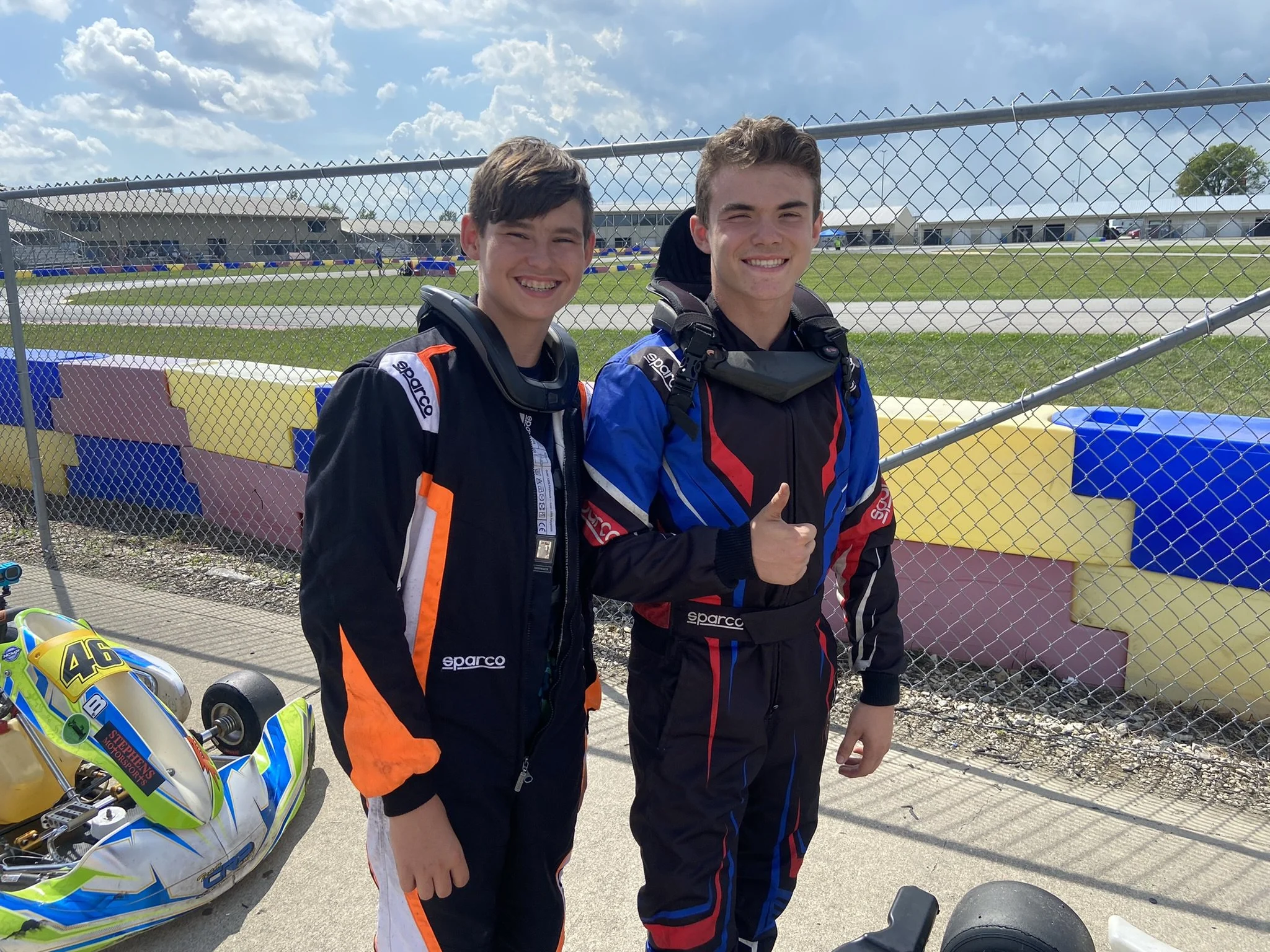 Two young race car drivers in racing suits standing next to go-karts outdoors at a race track, smiling, with a chain-link fence and grandstands in the background.