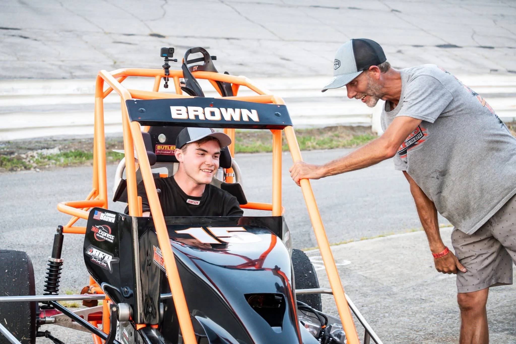 A young male race car driver in a black racing suit seated in a black and orange race car, talking to an older man in casual clothes and a gray cap, who is leaning on the car's orange frame, outdoors on a paved area.