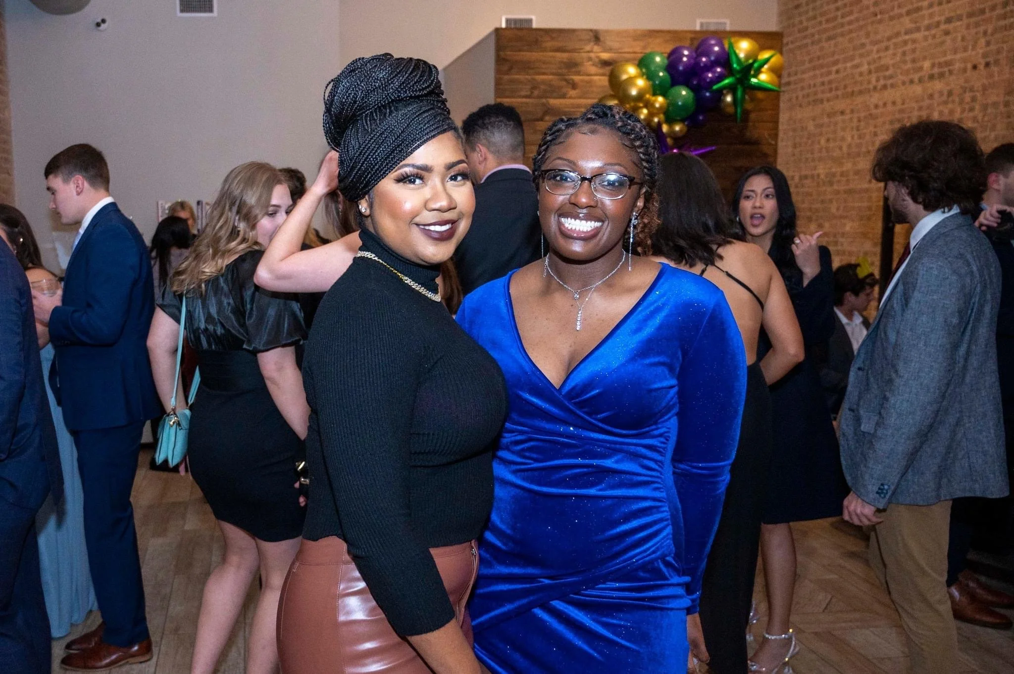 Two women at a social event, one wearing a black top and a head wrap, the other wearing a blue dress with jewelry, smiling at the camera. In the background, several people are mingling and talking.