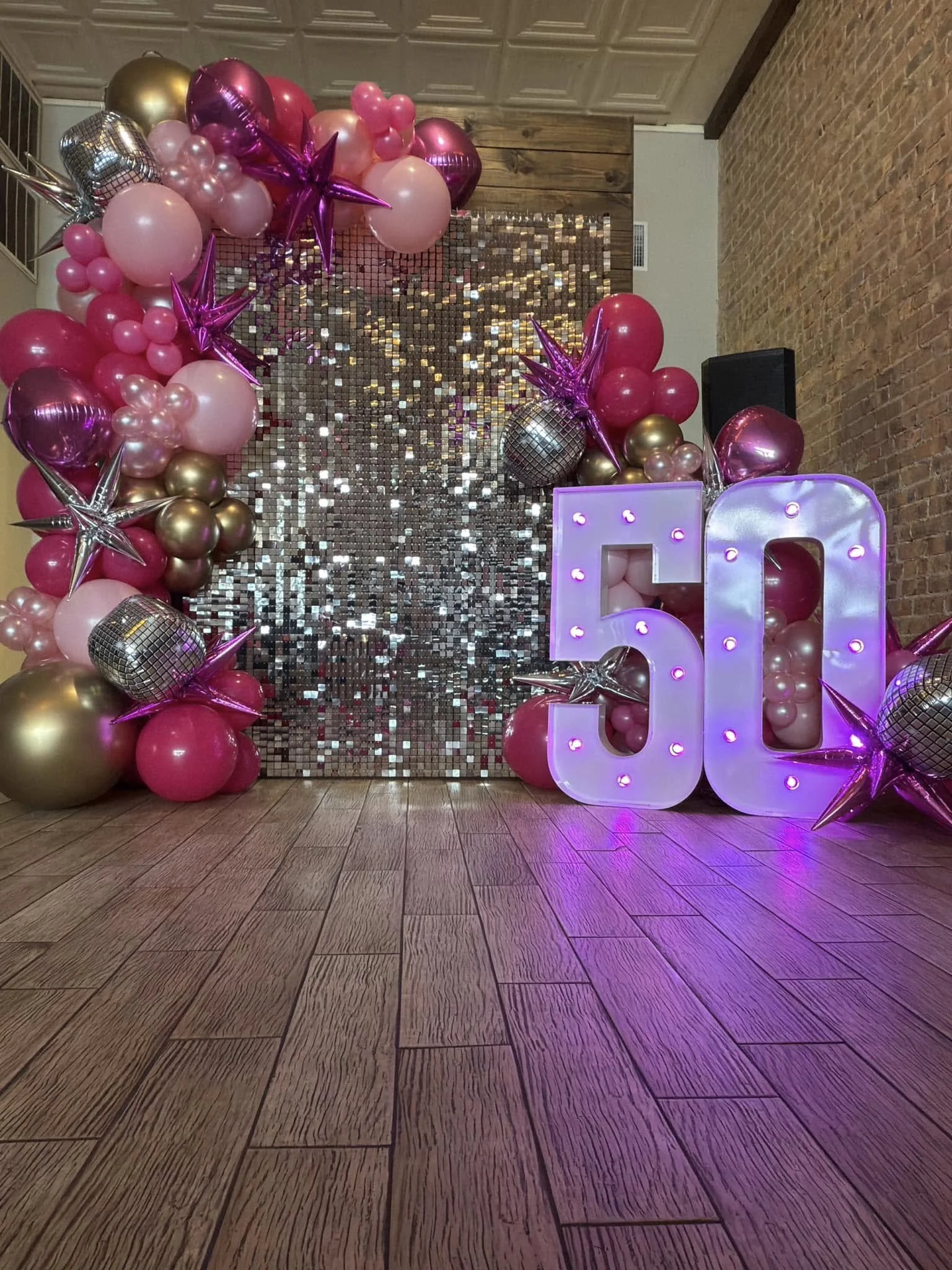 Pink, gold, and silver balloons arranged around a shiny sequin backdrop with large illuminated numbers '50' in front.