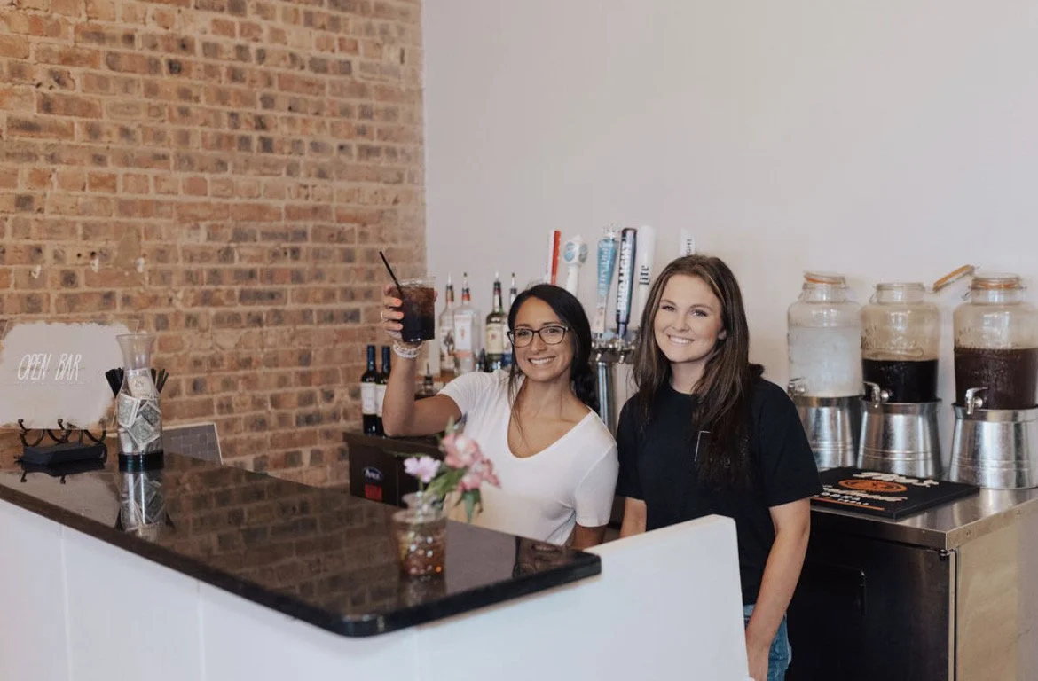 Two women working behind a bar, one holding a drink and smiling at the camera.