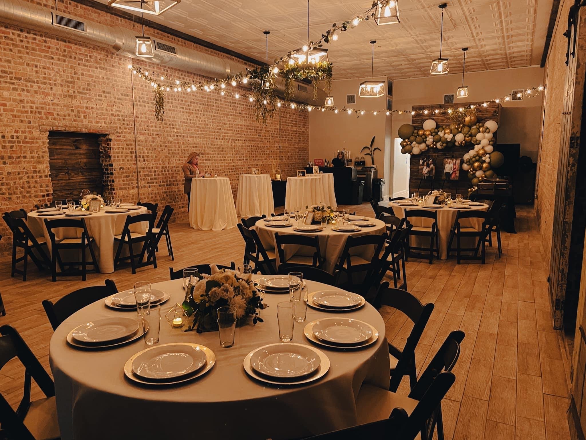 Round banquet tables with white tablecloths, set with white plates, glasses, and silverware, arranged in a decorated event space featuring brick walls, wooden flooring, string lights, and balloon arrangements.
